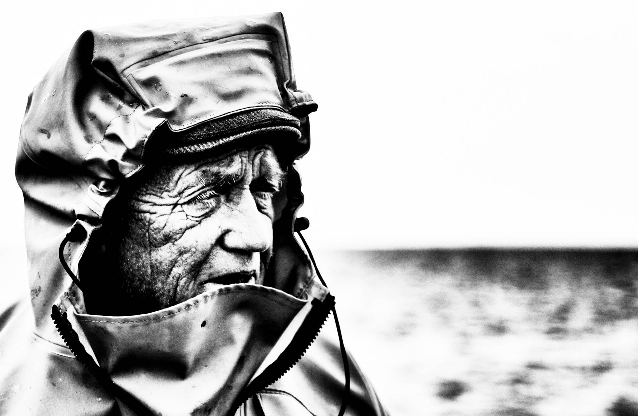 Elderly man in raincoat gazing at sea, black-and-white portrait, highlighting weathered features and ocean background.