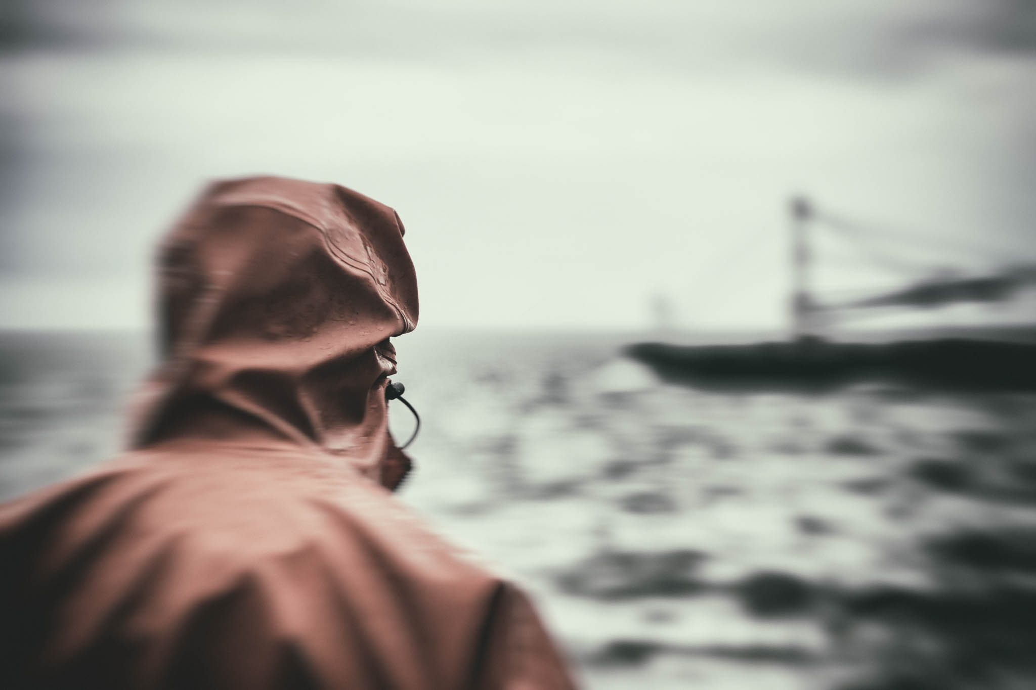 Person in raincoat gazes at blurred sea and distant ship under overcast sky; maritime adventure, moody atmosphere.