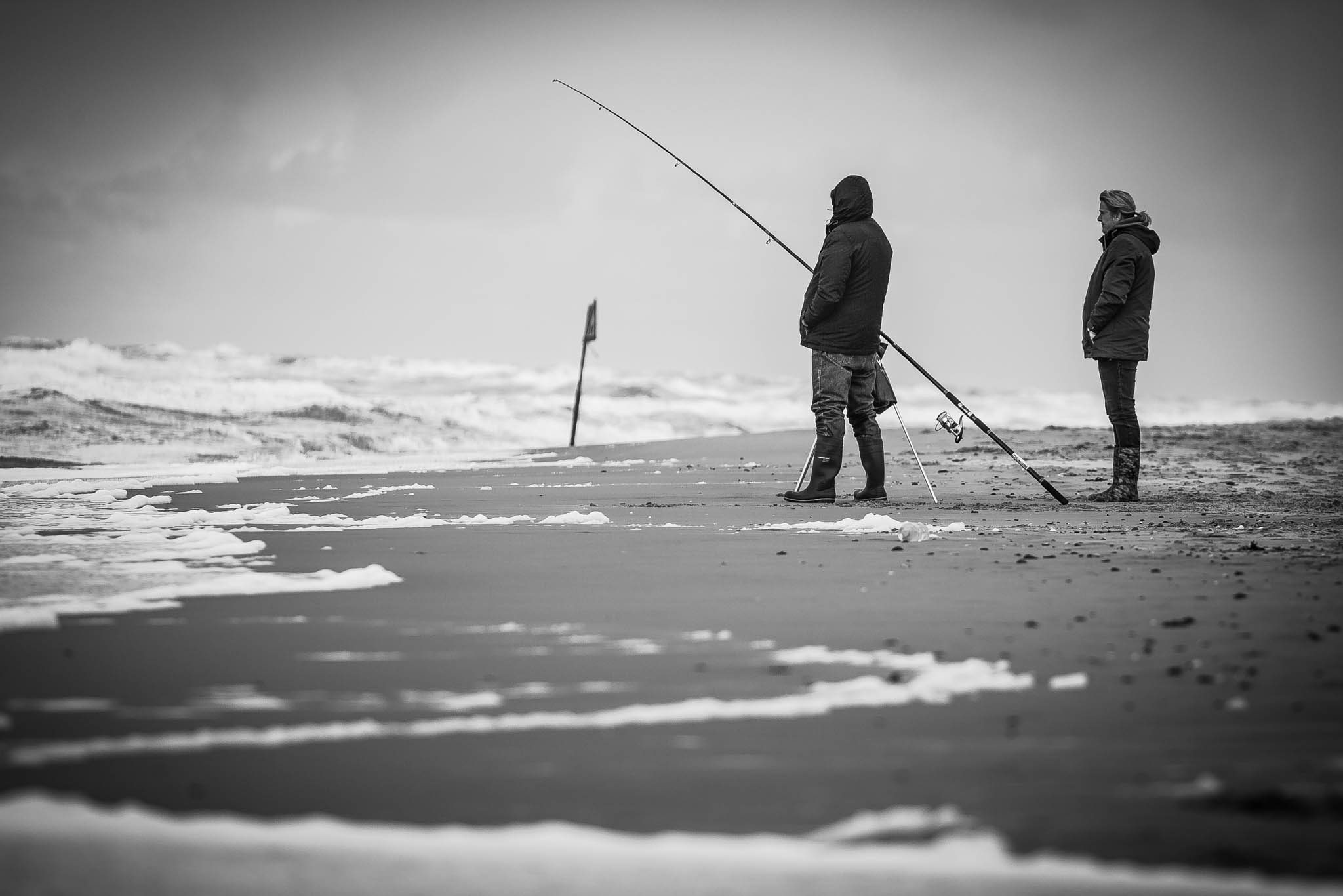Two people beach fishing on a windy, cloudy day, standing on the shore with ocean waves in the distance.