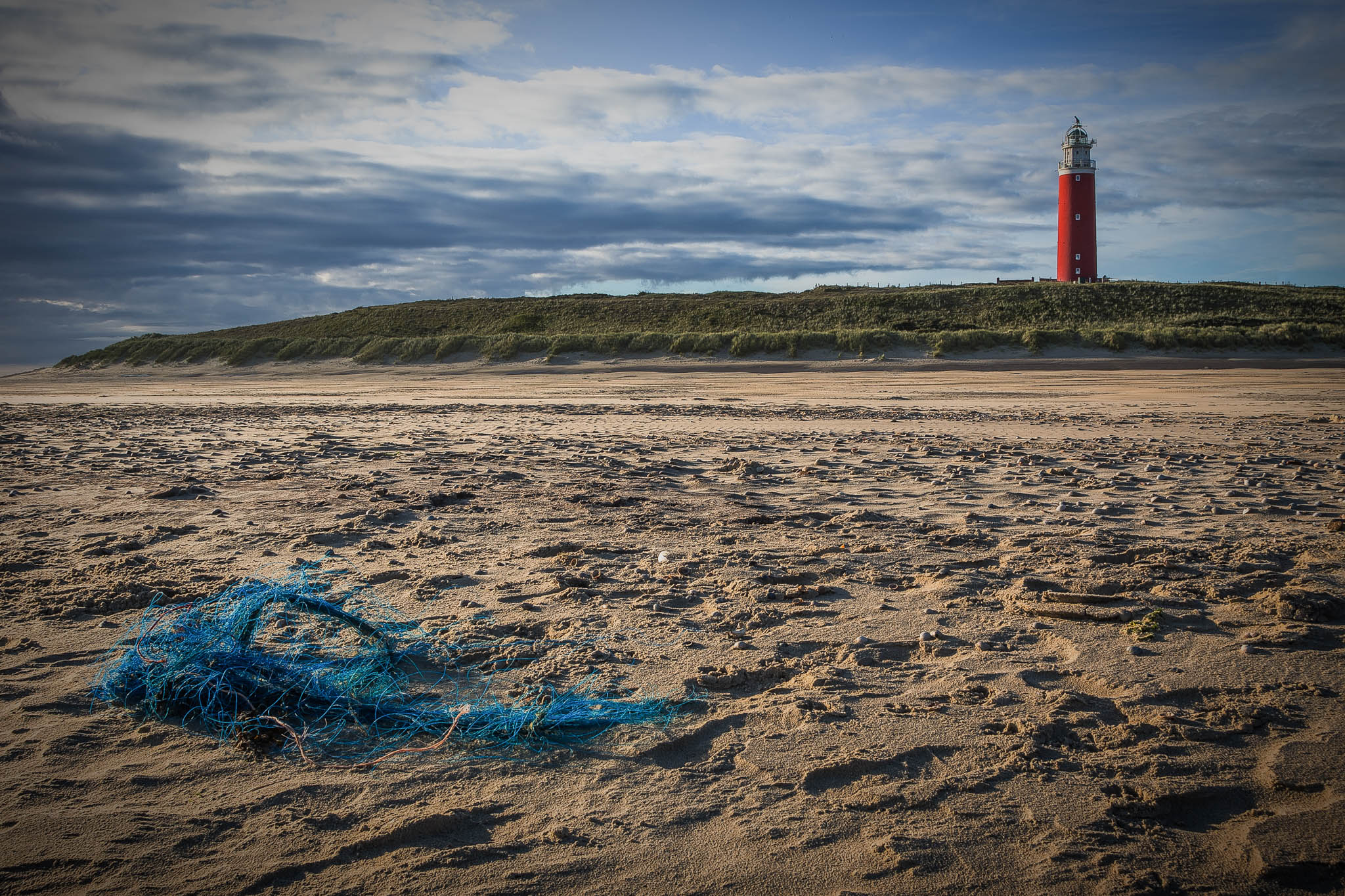 Blue fishing net on sandy beach with red lighthouse in background under cloudy sky.