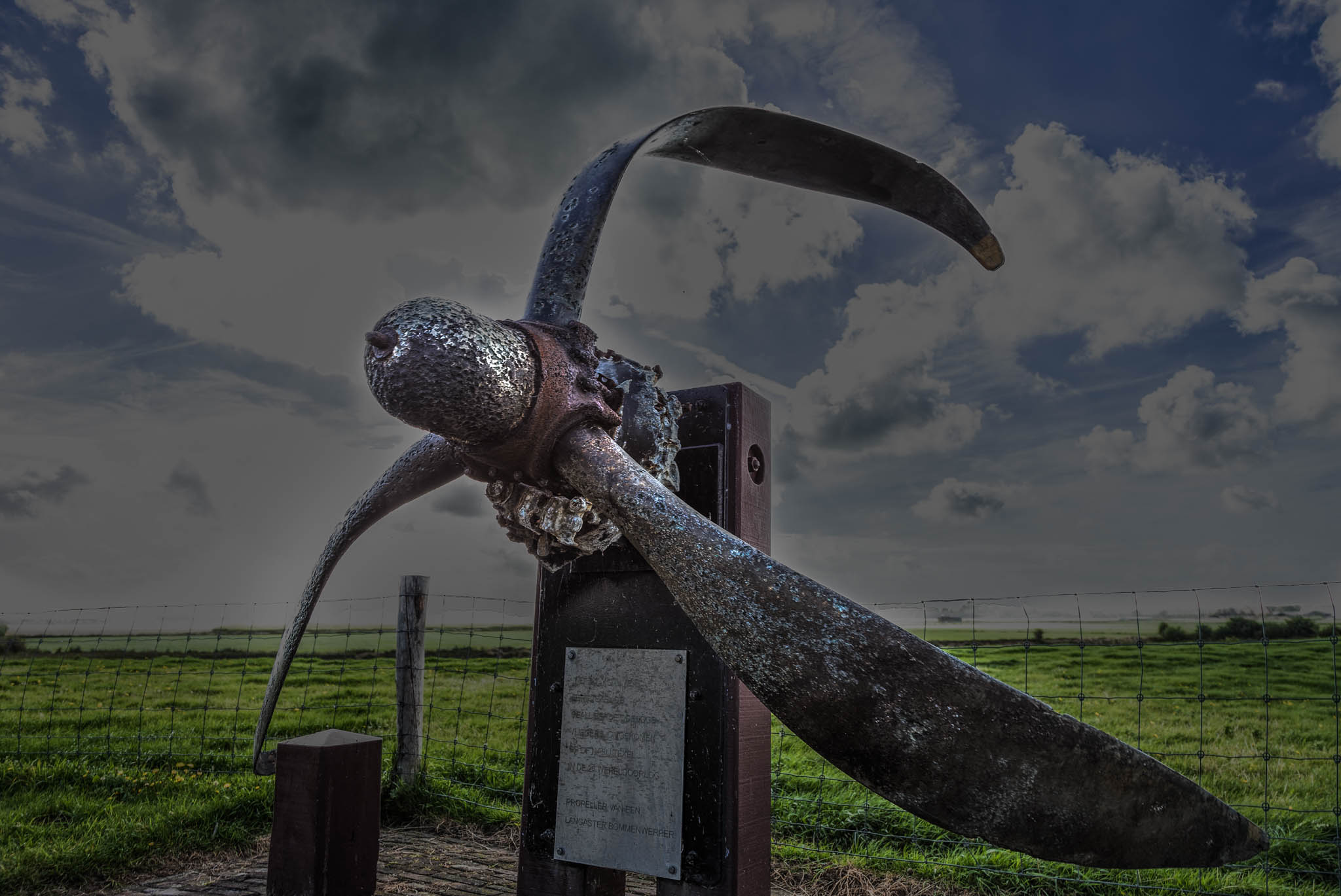 Rusty aircraft propeller monument against a cloudy sky in an open grassy field.
