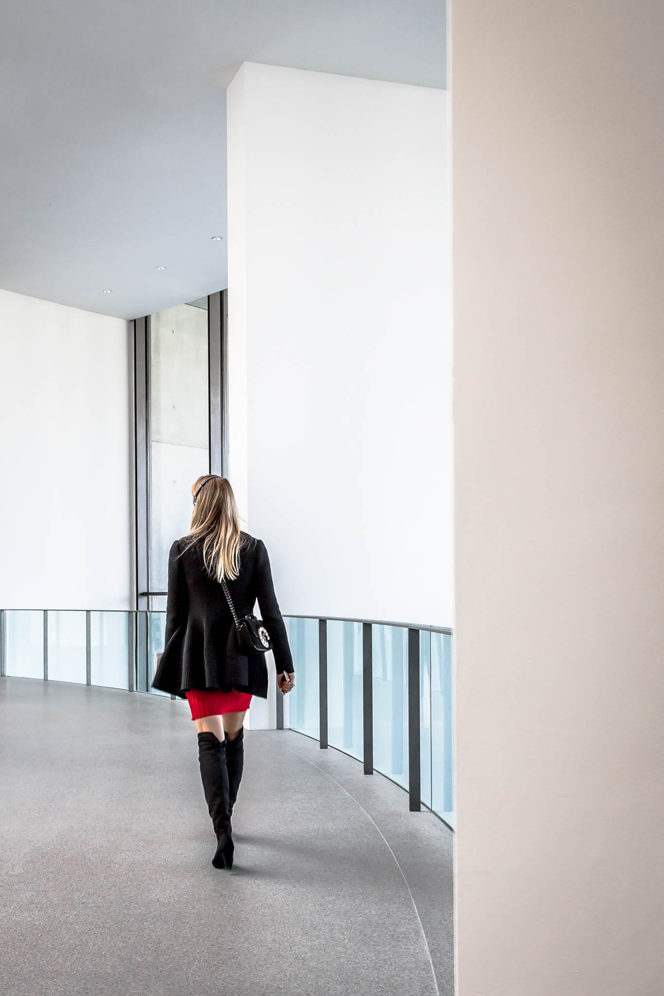 Woman in black coat and red dress walking in modern, bright hallway with glass railing.