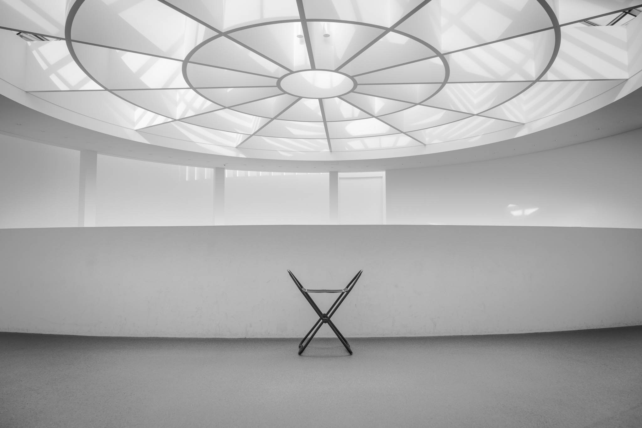 Minimalistic interior with a geometric ceiling and a lone chair in a bright, open space.