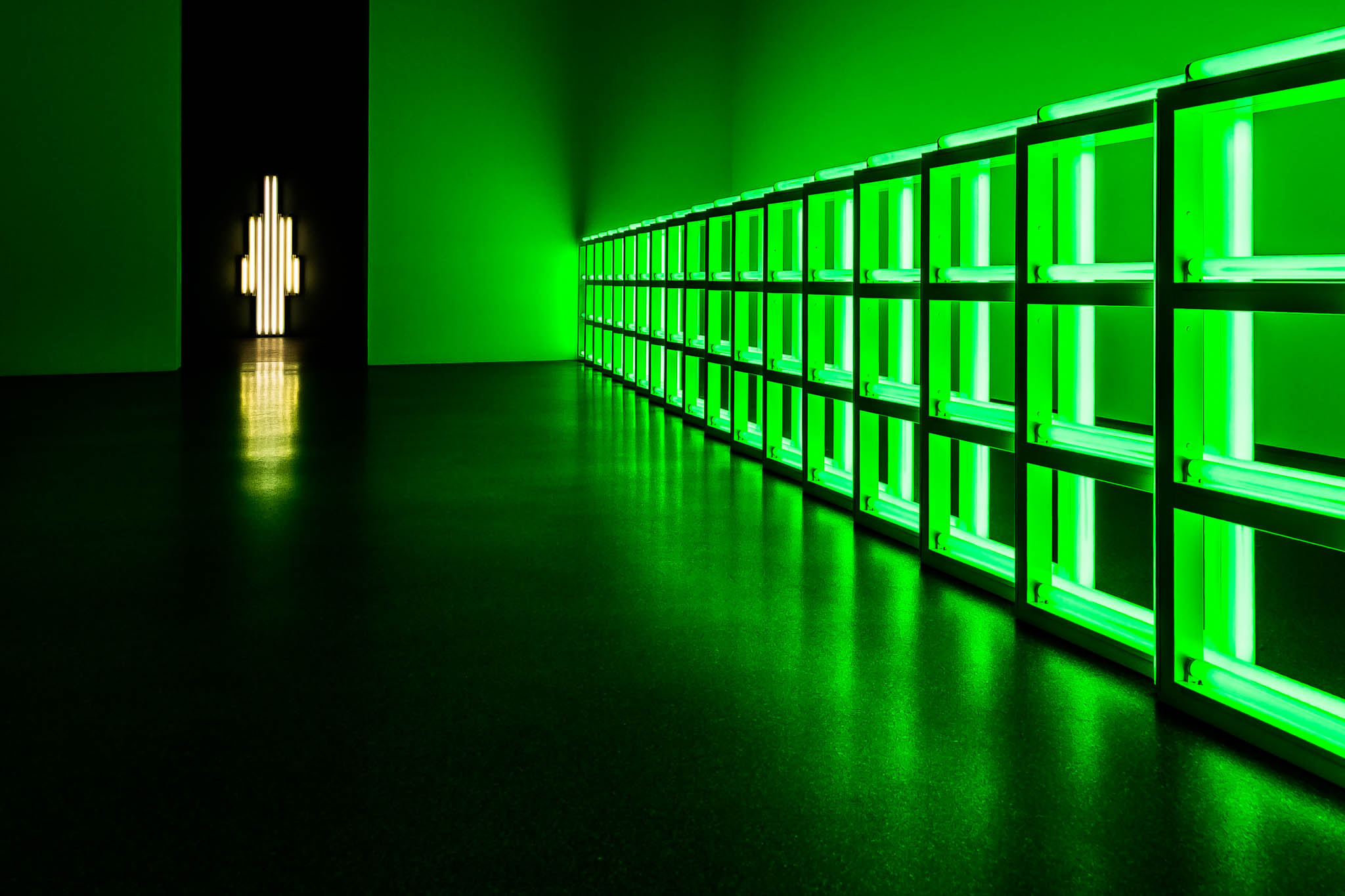 A row of green neon lights lines a gallery wall, with a distant glowing white light in a dark doorway.