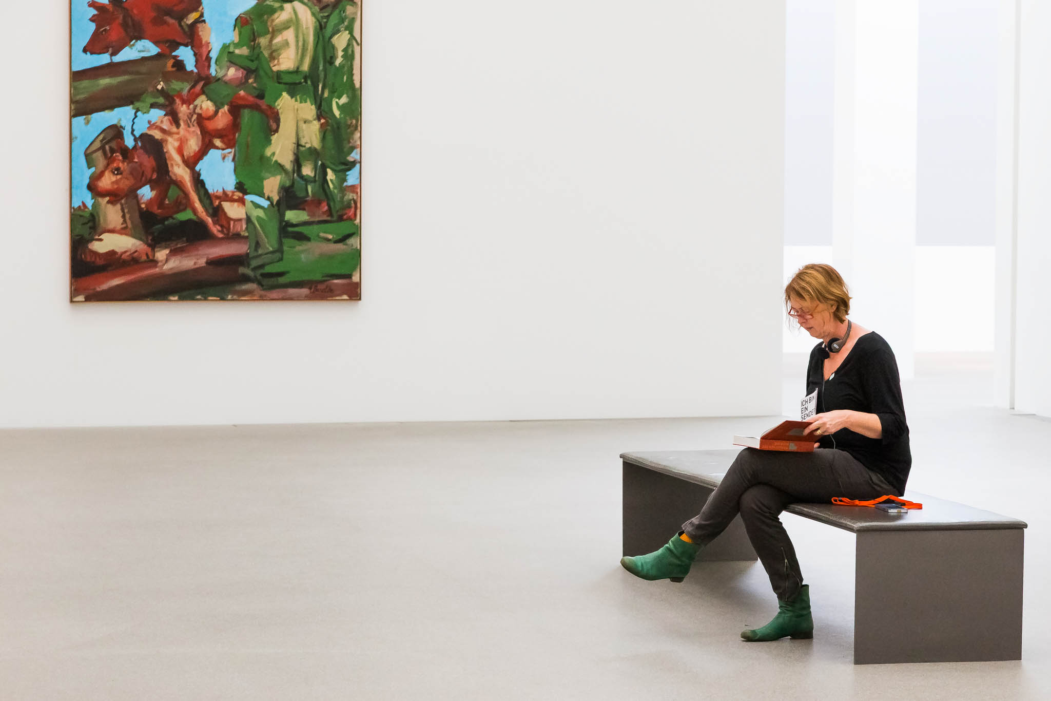 Woman seated on bench reading a book in modern art gallery, abstract painting on white wall.