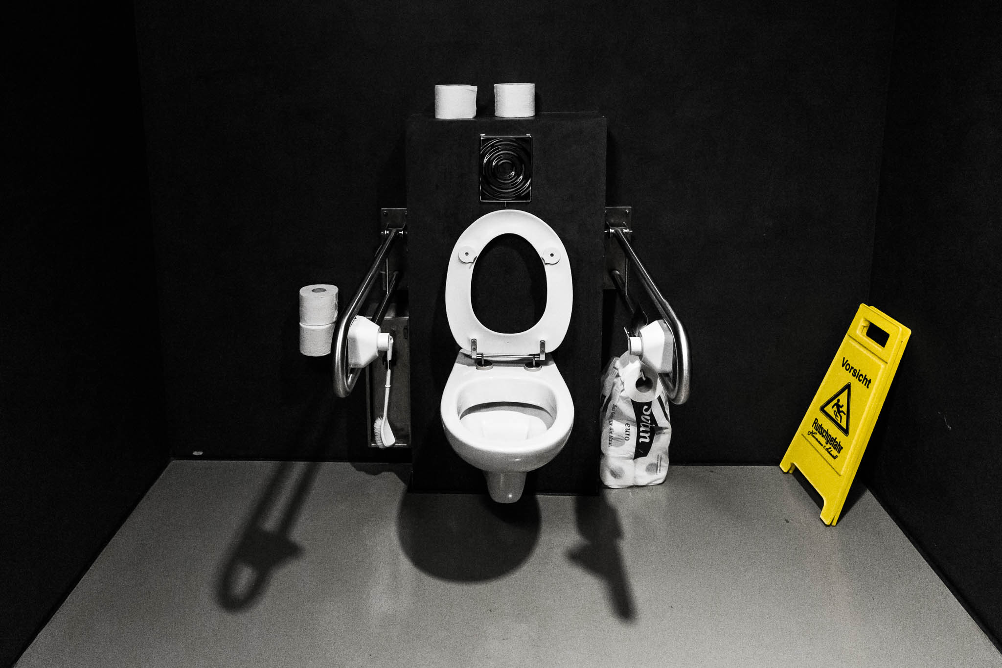 Accessible restroom with grab bars, toilet paper, and caution sign on a dark background.