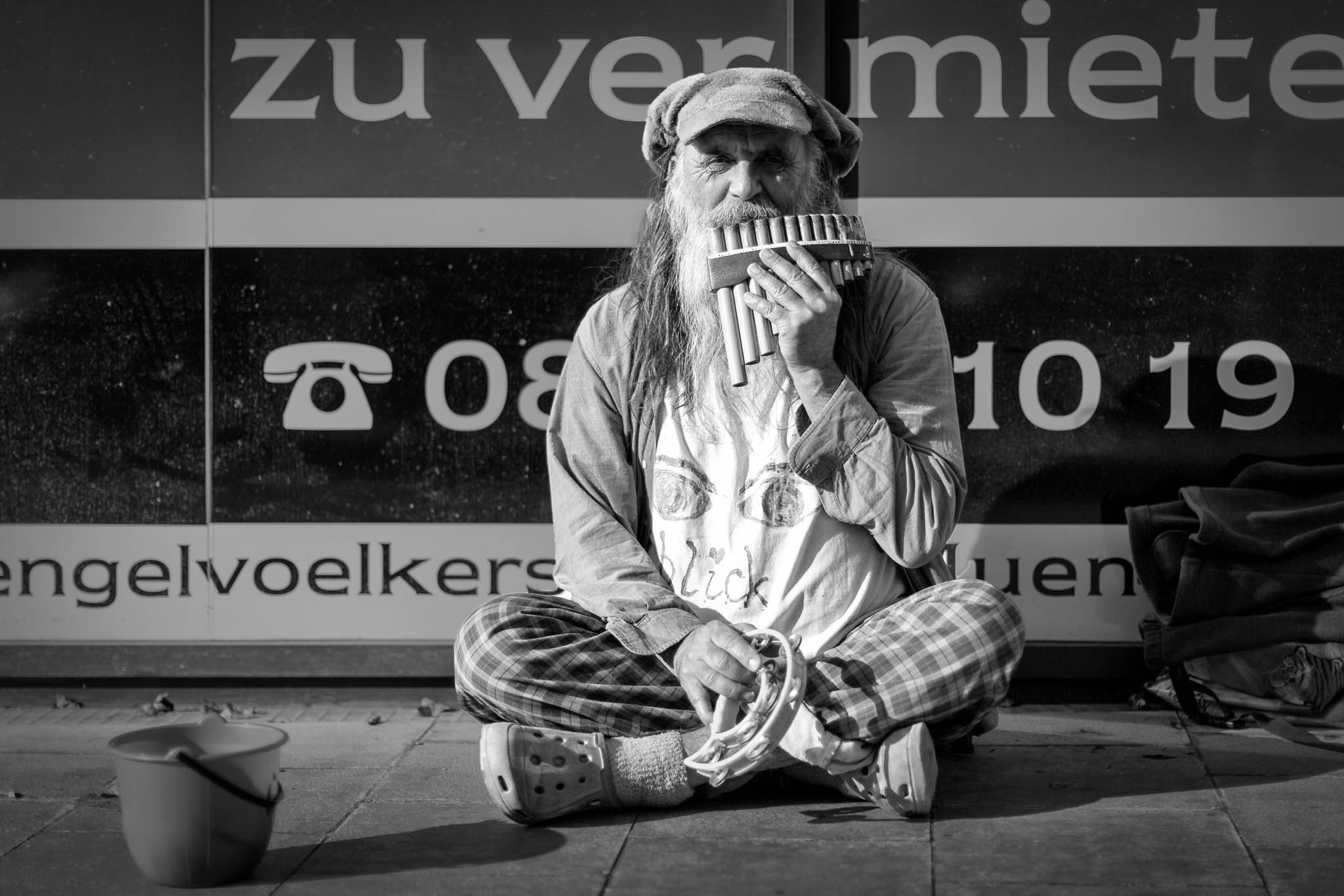 Street musician playing pan flute, sitting cross-legged with tambourine in hand, beside a zu vermieten (for rent) sign.