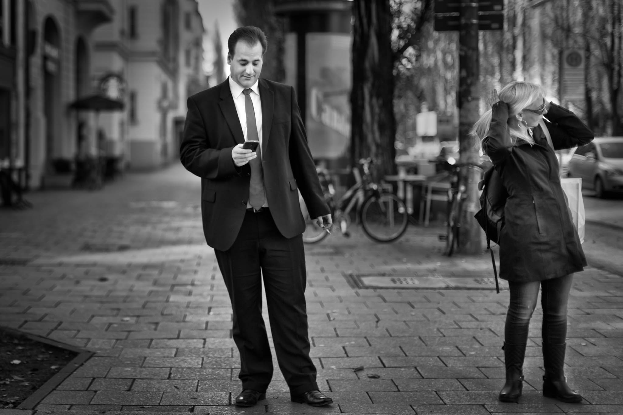 Man in a suit checks phone, while woman adjusts hair on a city sidewalk; bicycles and cars in the background.
