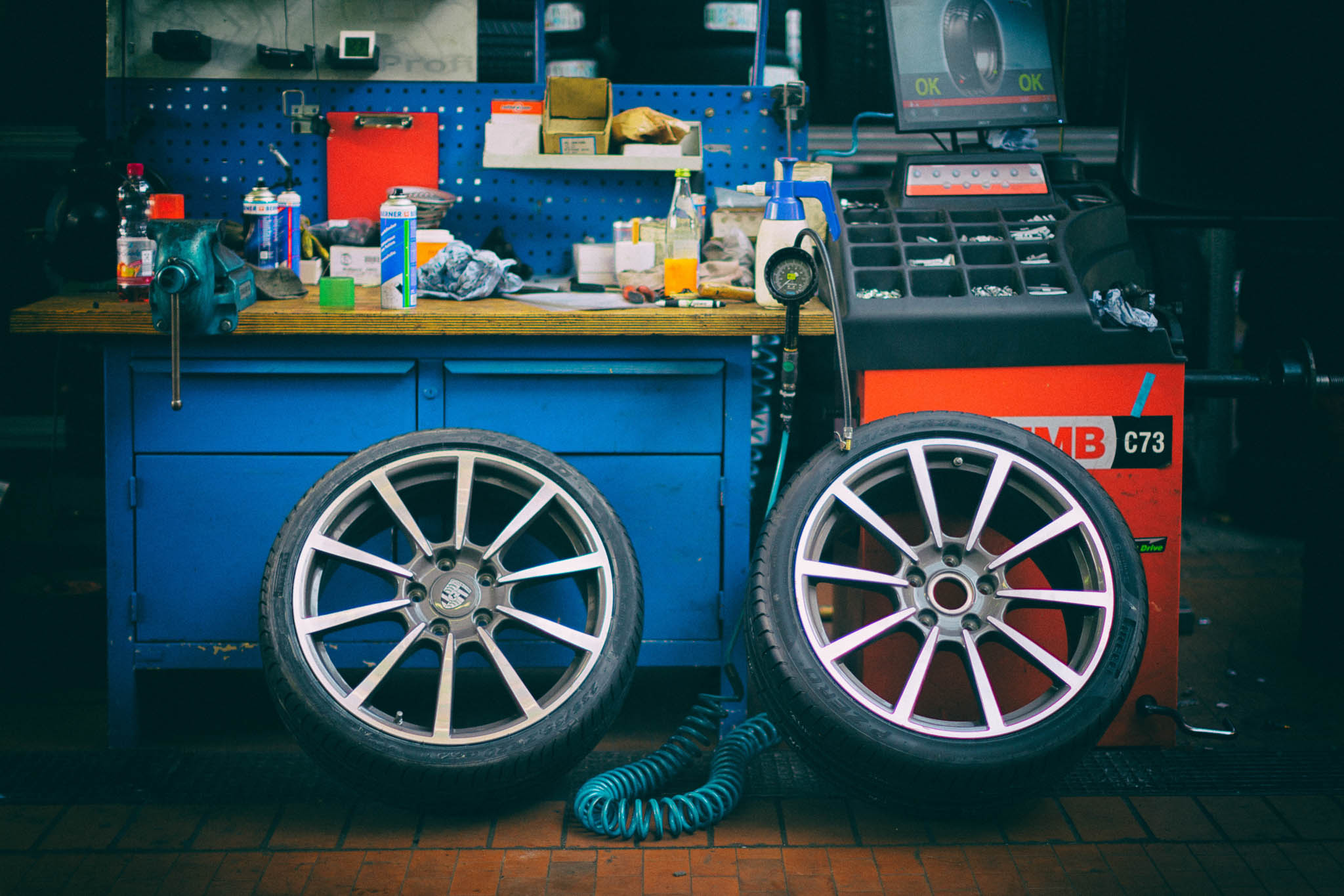 Car workshop with two alloy wheels, workbench tools, and a tire balancing machine in the background.