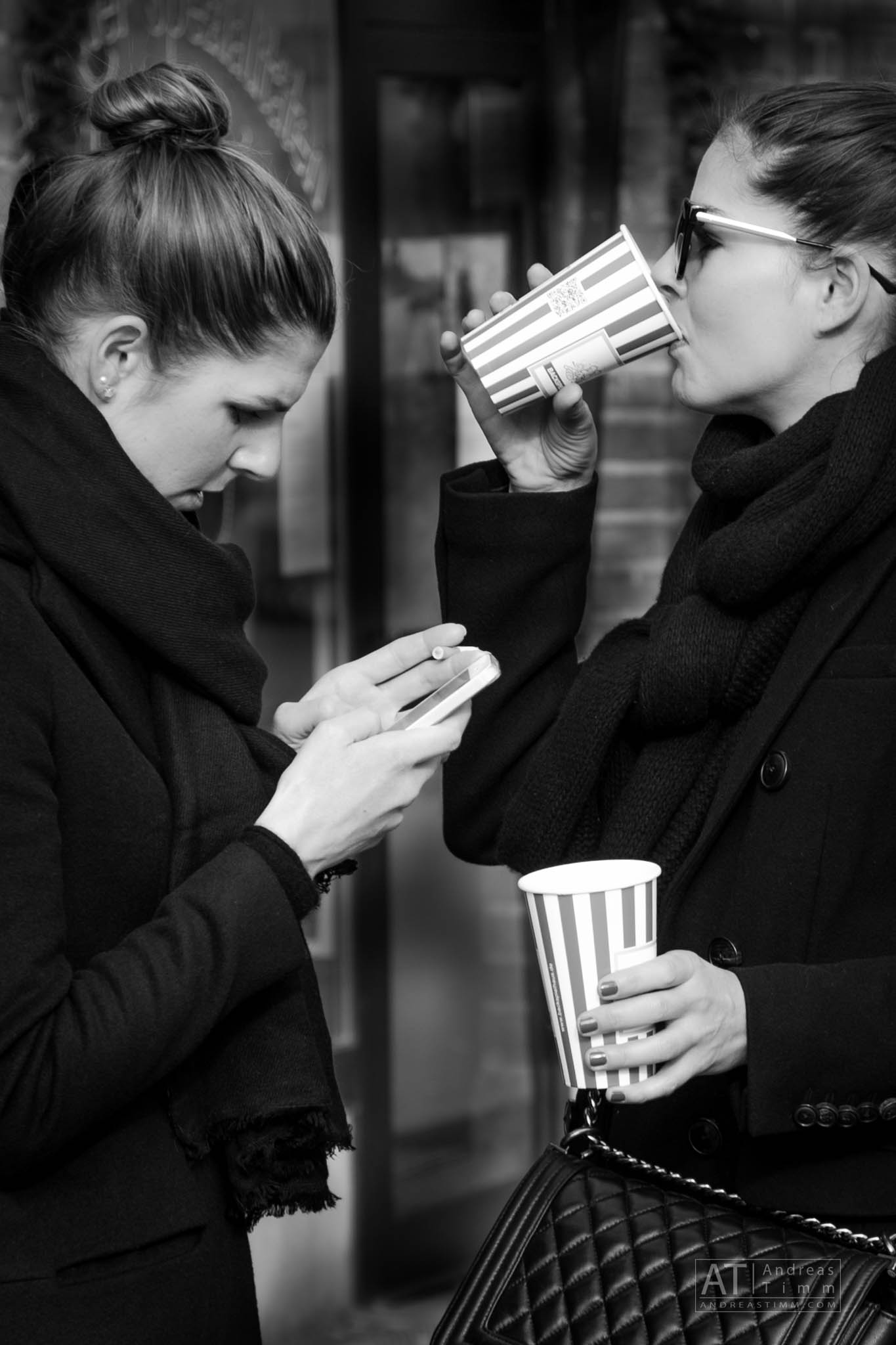 Two women in black coats, one using a smartphone, the other drinking from a striped coffee cup.