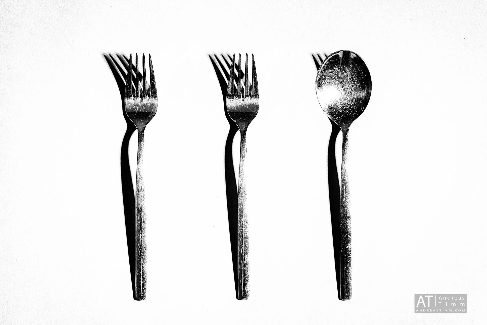 Three metal utensils including two forks and a spoon casting long shadows on a white surface.