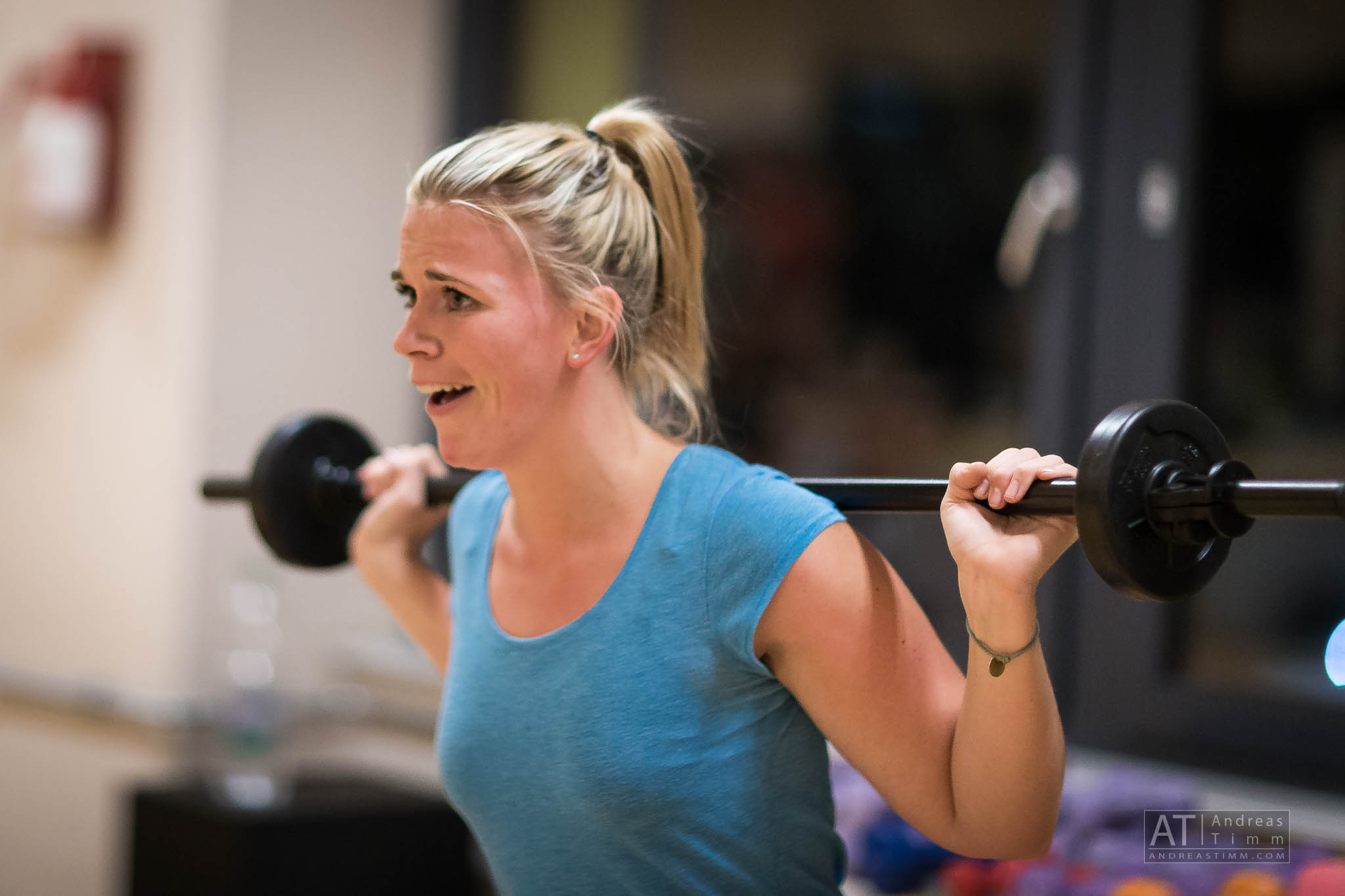 Woman lifting a barbell in a gym, wearing a blue shirt, showing focused expression during workout.