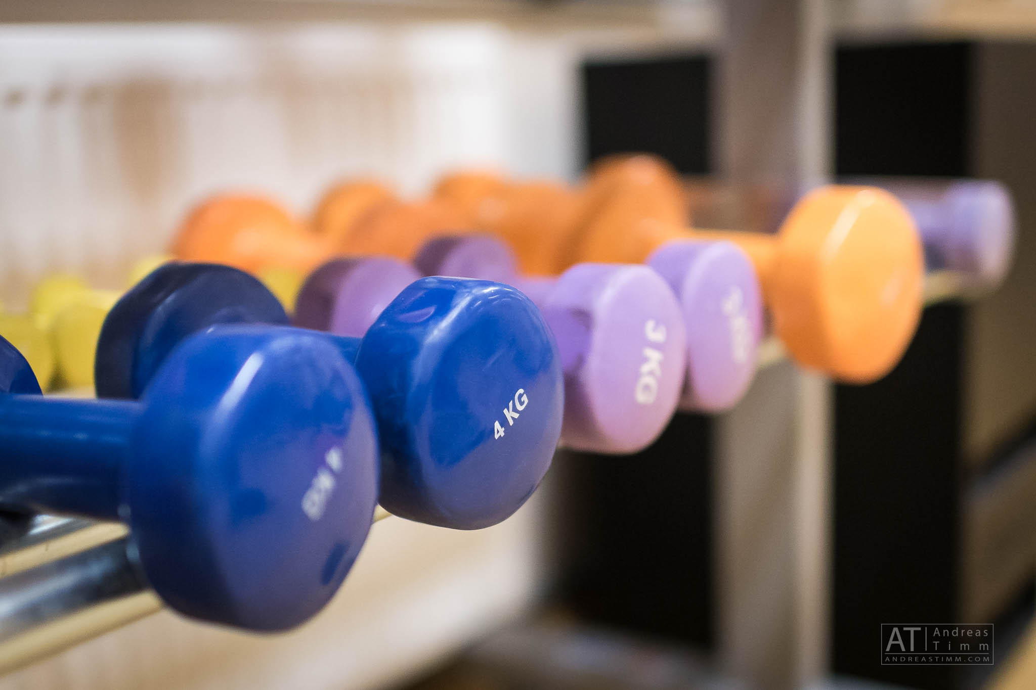 Colorful dumbbells arranged on a rack, including 4 kg blue and 3 kg purple weights, in a gym setting.