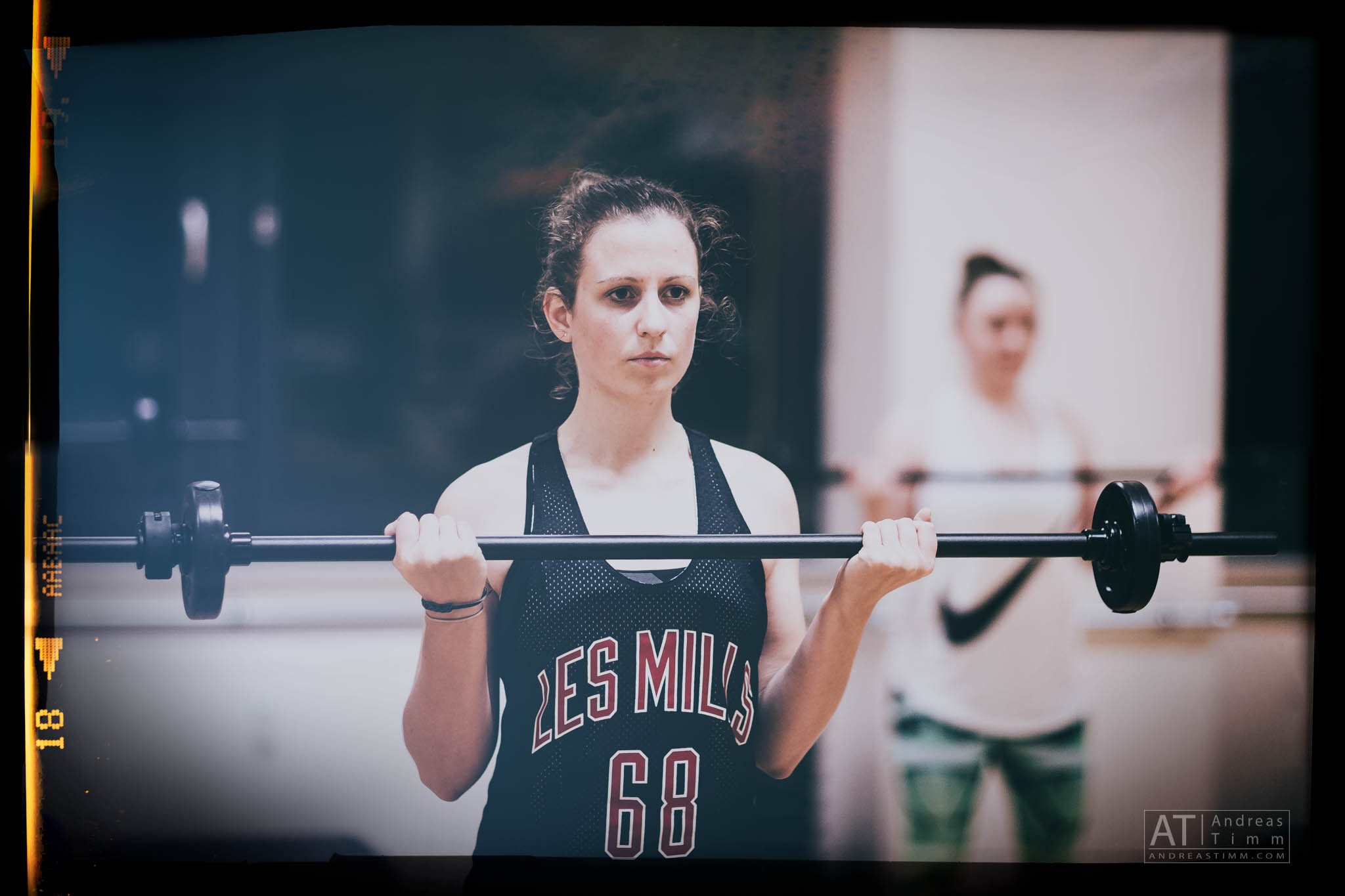 Woman lifting barbell in gym, focused expression, wearing Les Mills tank top. Exercise class in background.