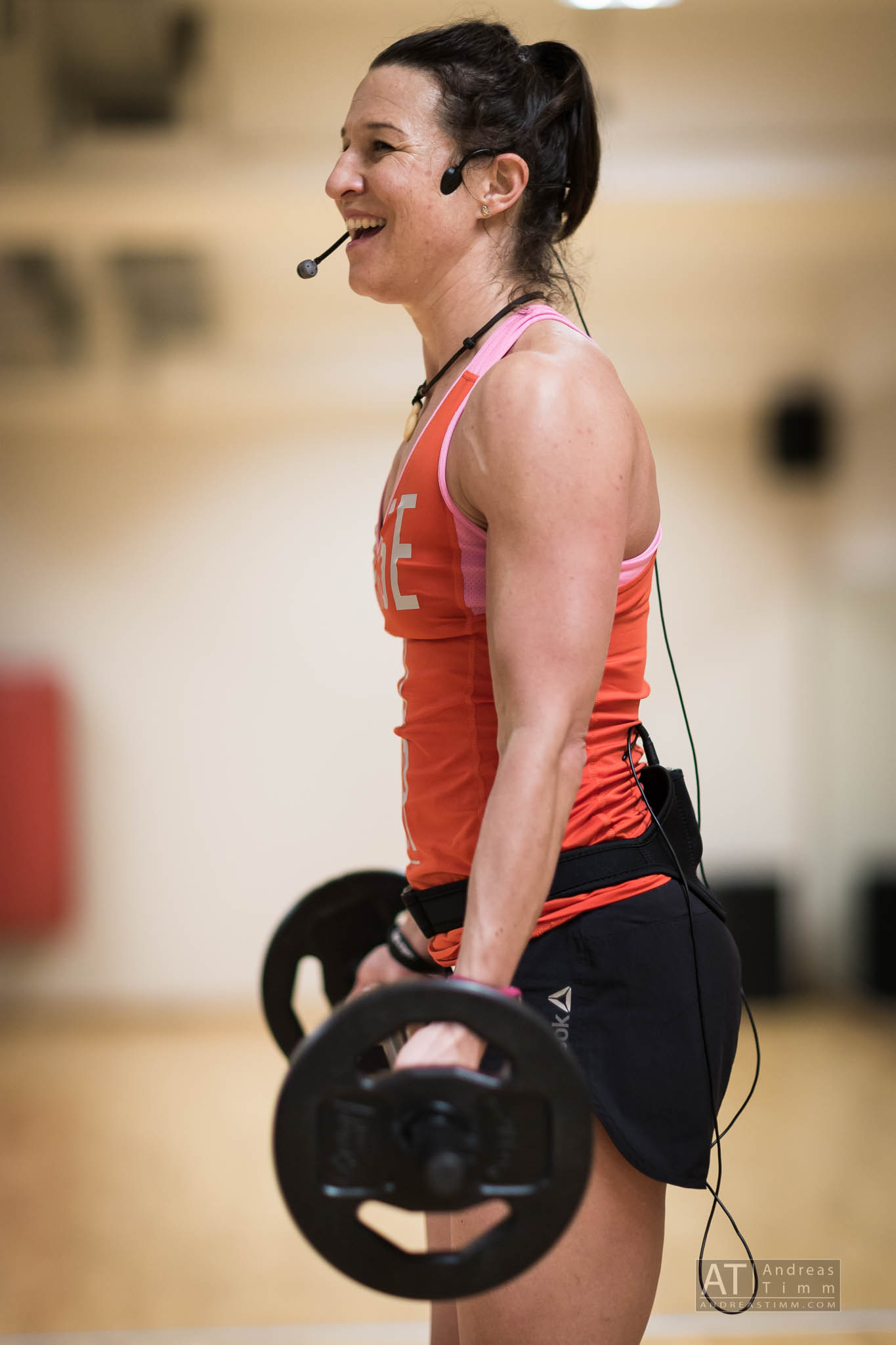 Fitness instructor with headset and dumbbell, smiling in gym, wearing orange tank top and black shorts.