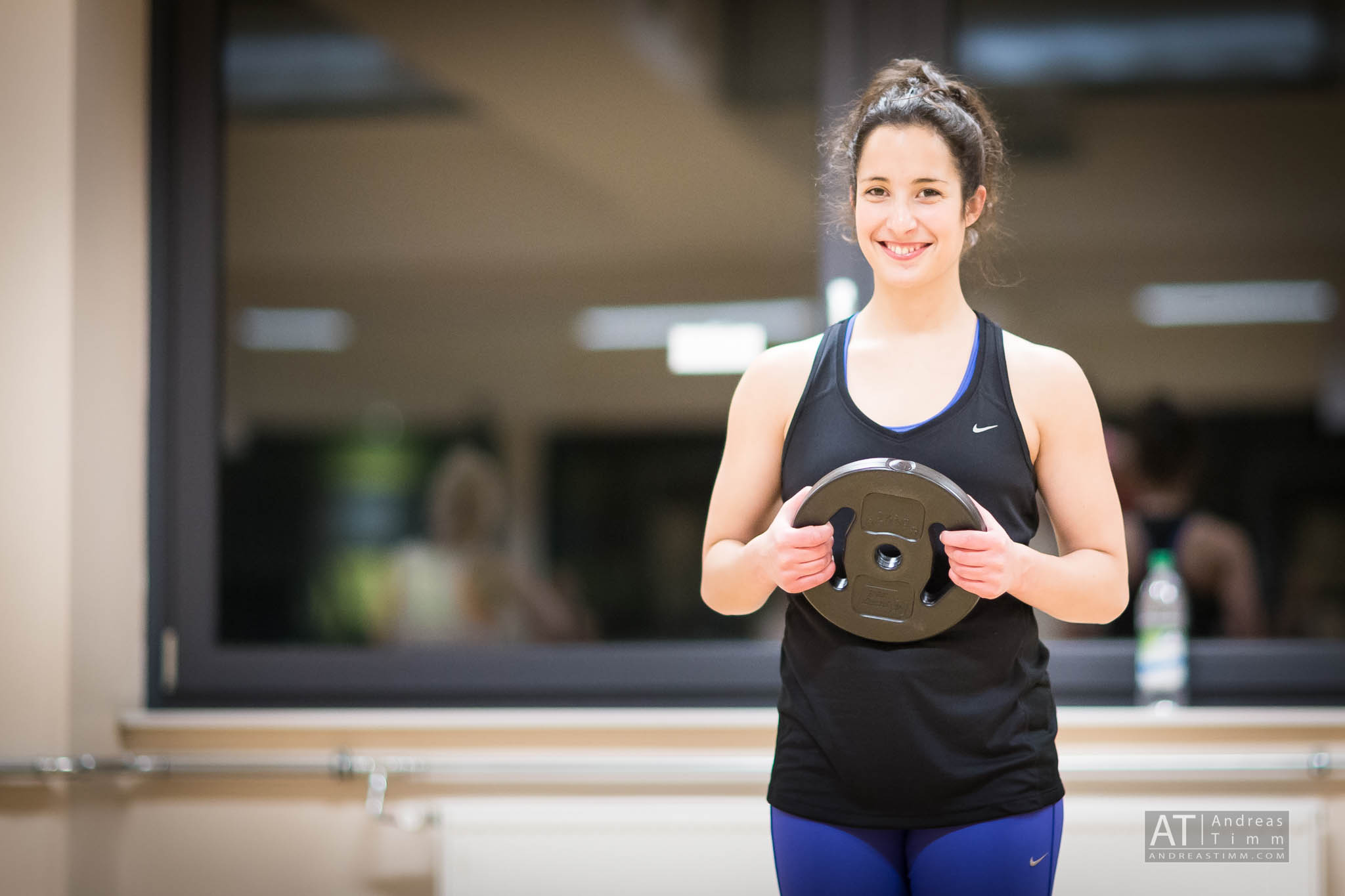 Woman in gym holding a weight plate, smiling, wearing a black tank top and blue leggings.