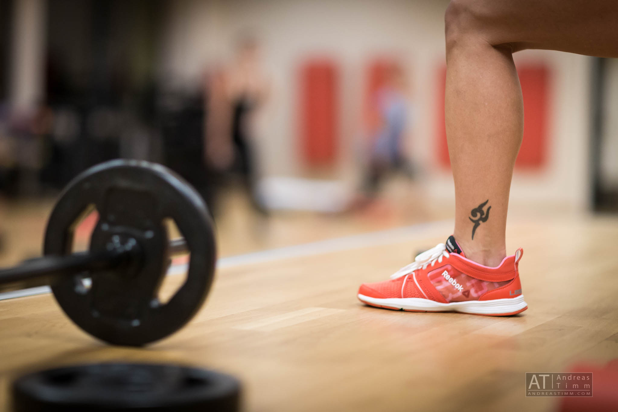 Leg in red Reebok shoe and tattoo next to barbell in gym.