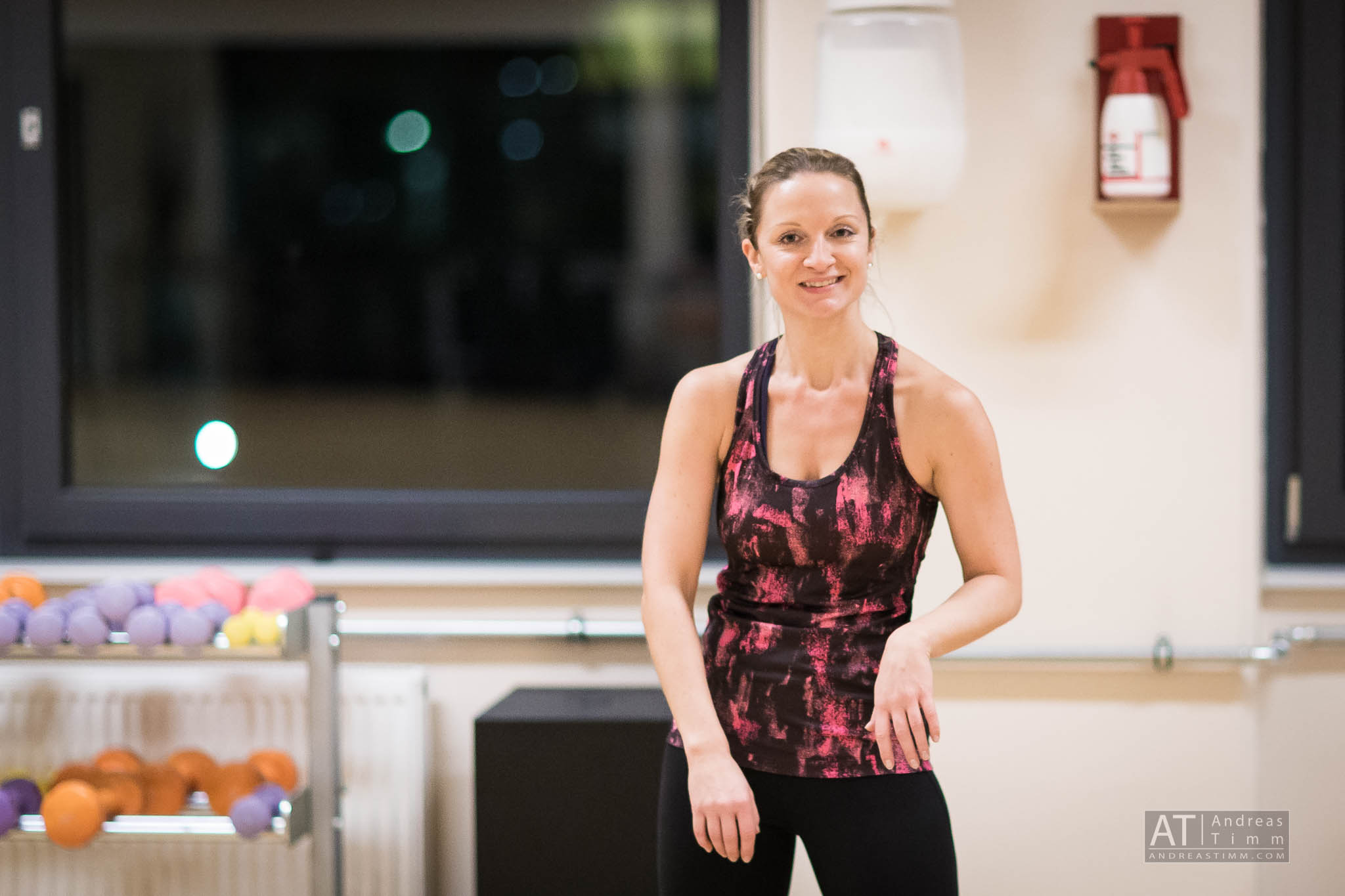 Woman in a gym wearing a pink and black tank top, smiling near exercise equipment and a window.