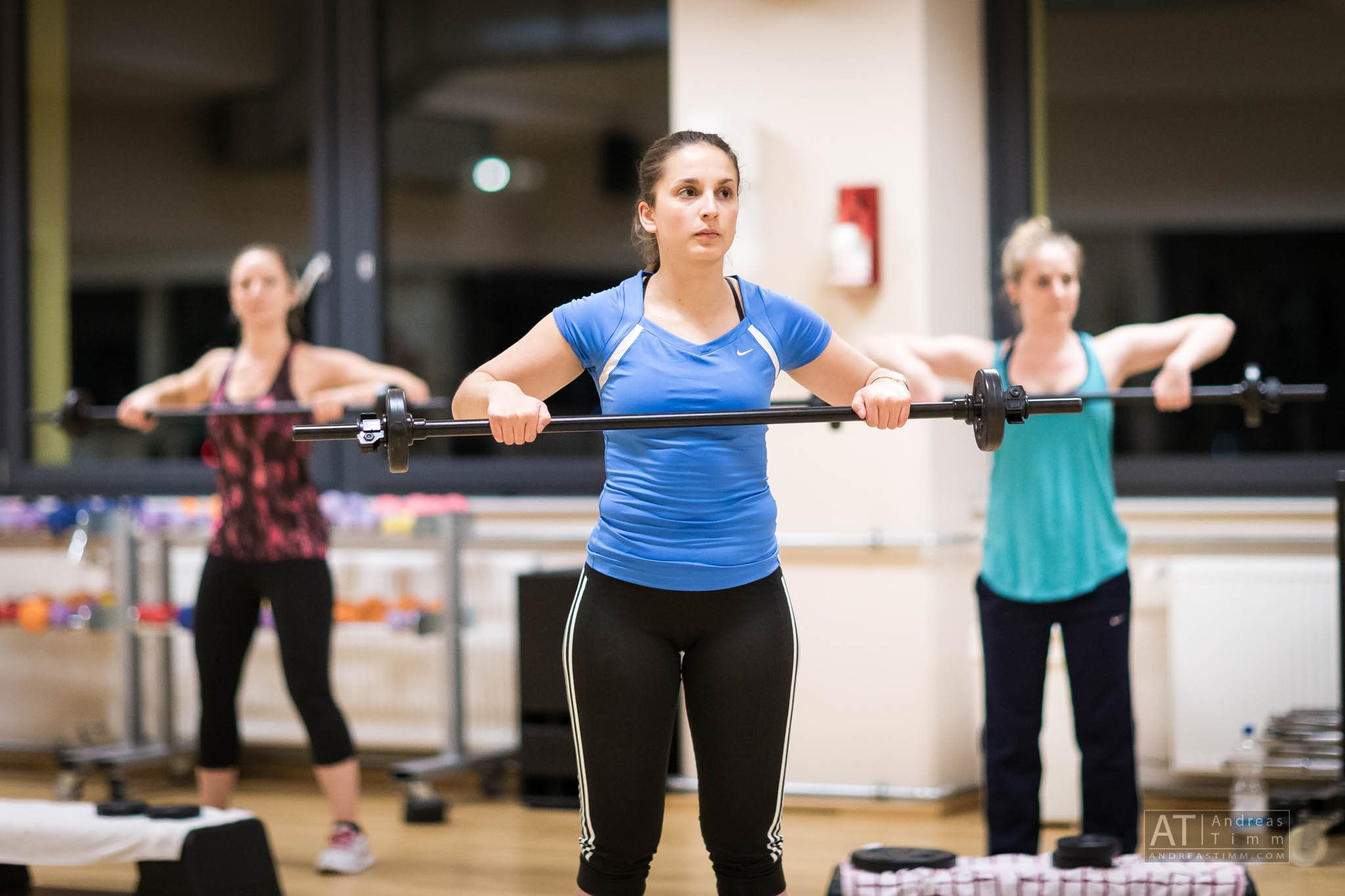 Women lifting barbells in a fitness class, focusing on strength training in a bright gym environment.