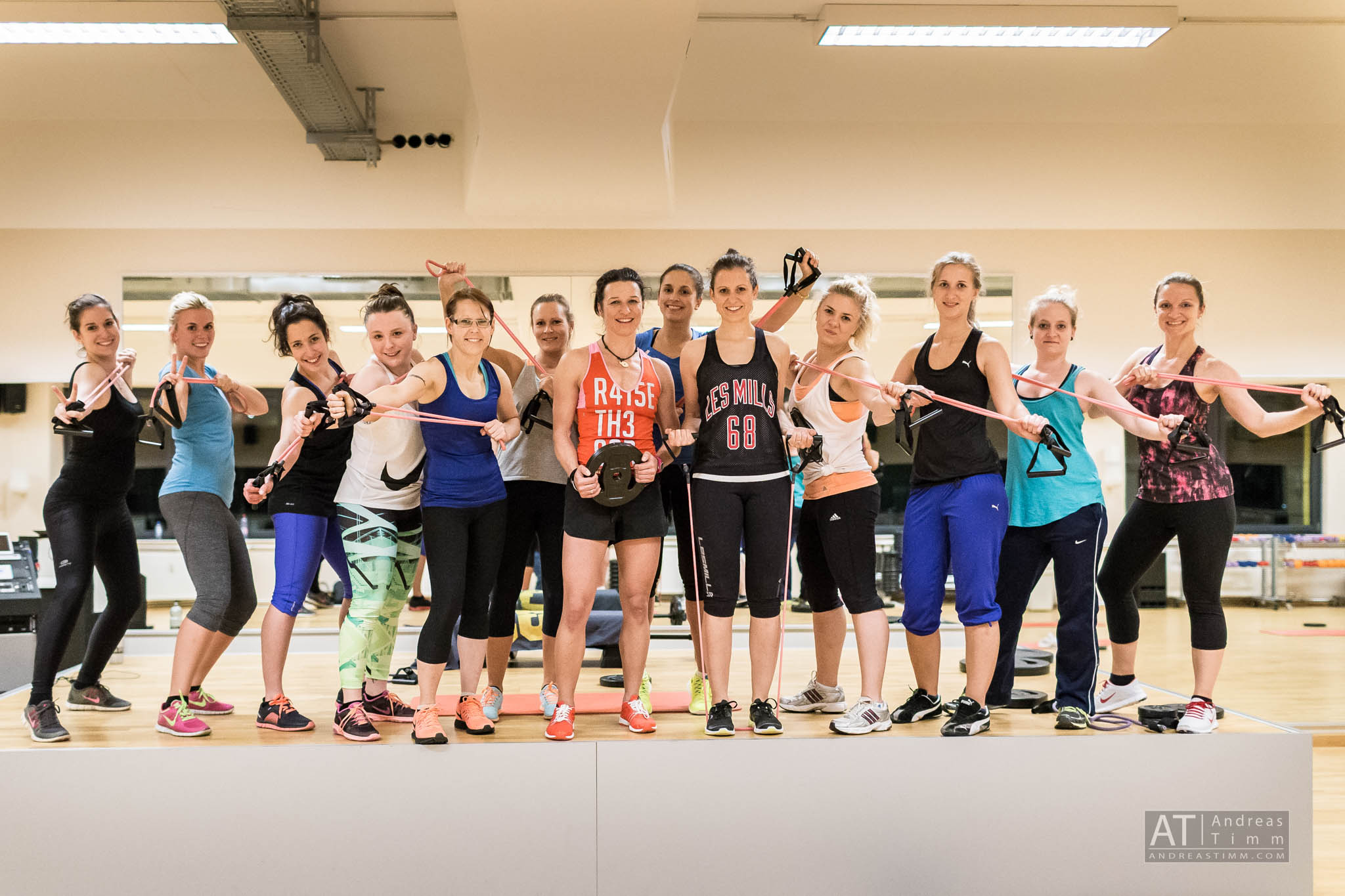 Group fitness class with women holding resistance bands, smiling in a gym studio.