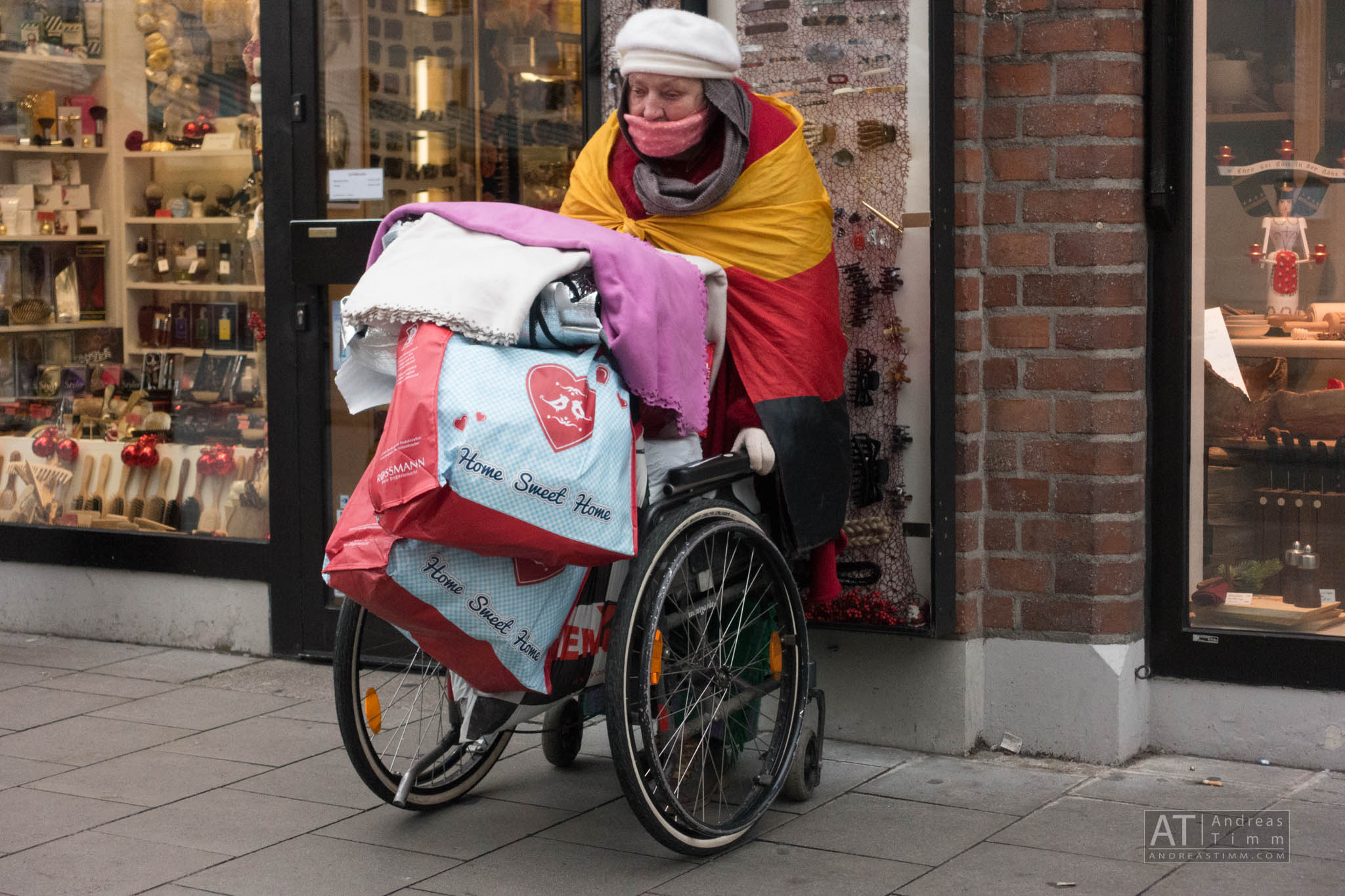 Person in wheelchair covered with colorful blankets and bags, outside a shop displaying various items, winter scene.