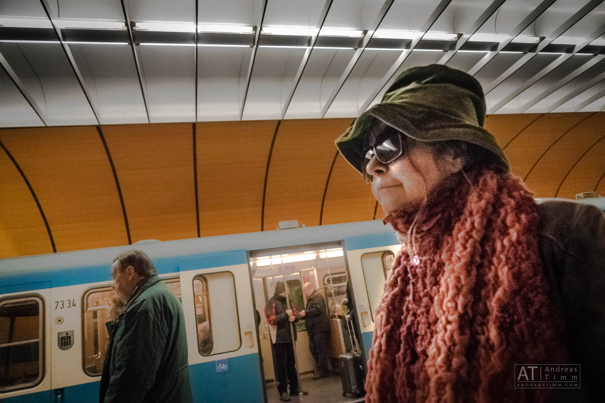 Woman in hat and scarf at a subway station, next to a blue train, with an orange ceiling overhead.