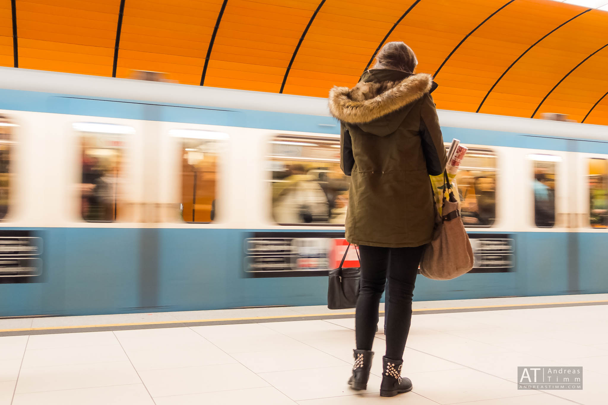 Woman in a winter coat waits as a blue subway train speeds past an orange underground station.