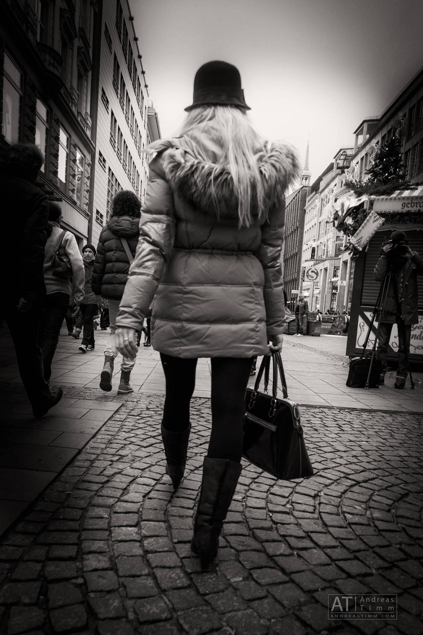 Woman in fur-trimmed coat and hat walking through a cobblestone street in a bustling city, carrying a large handbag.