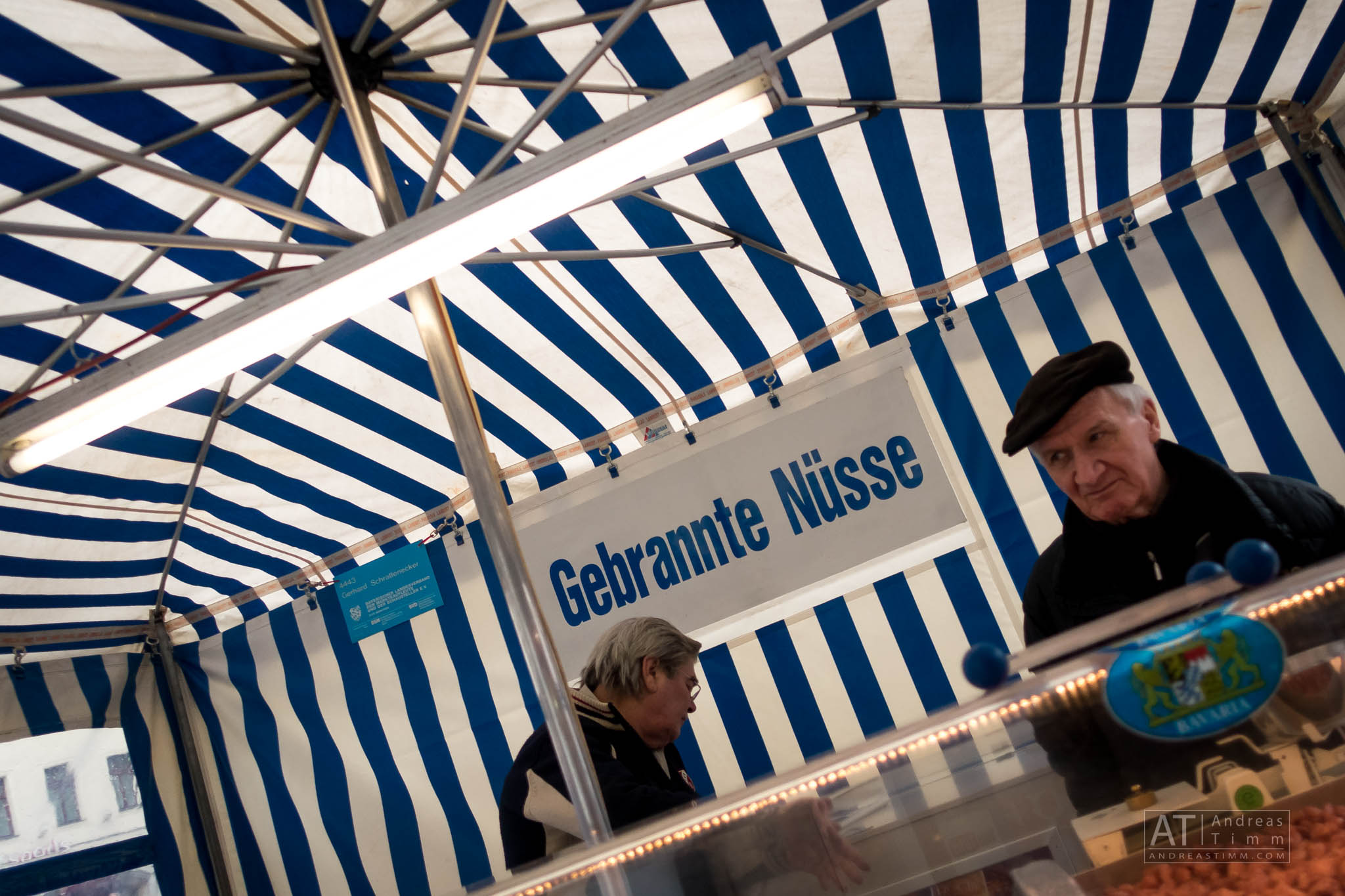 Market stall with blue-and-white striped canopy selling Gebrannte Nüsse (roasted nuts), man in black hat present.