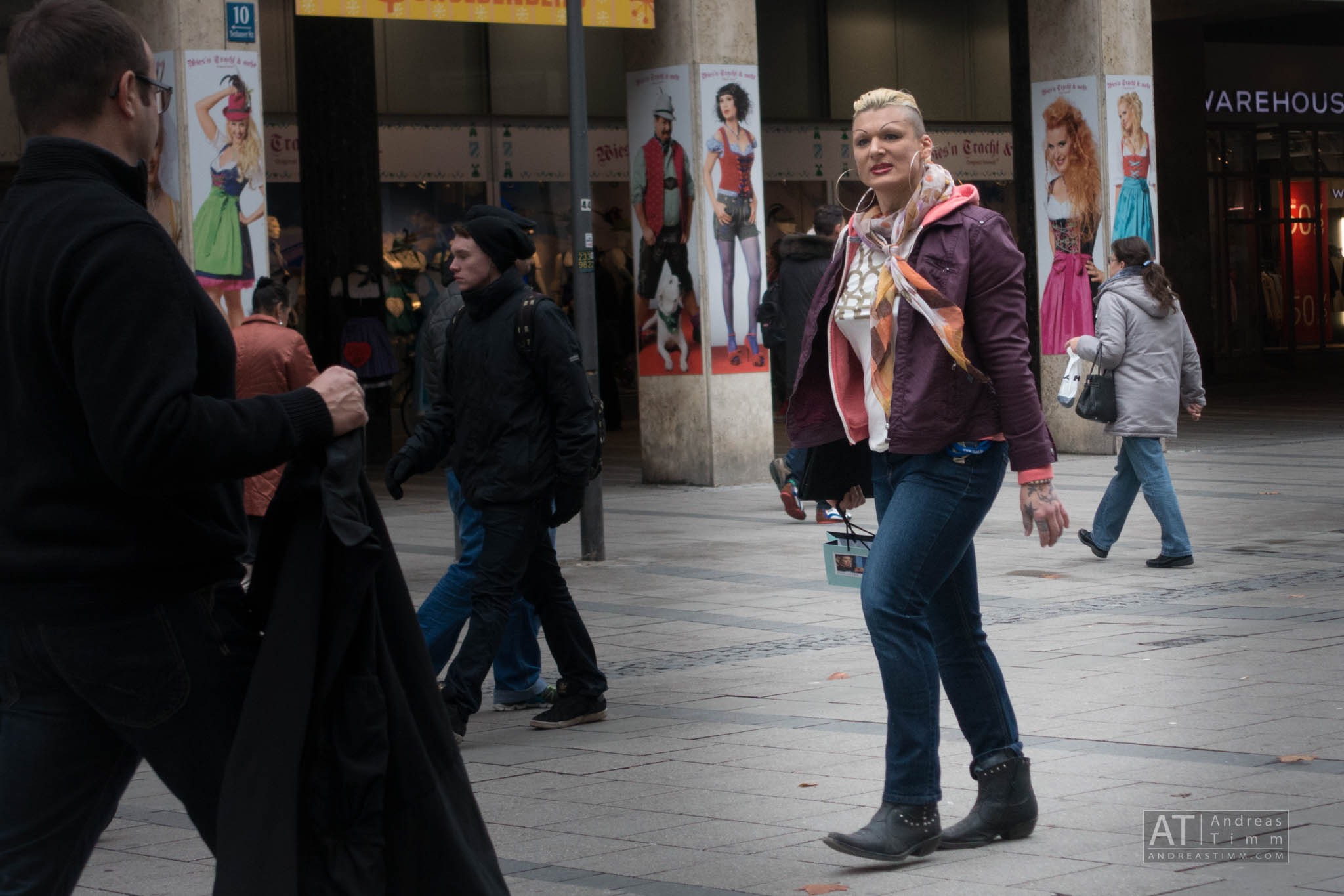 People walking on a busy street with shopping bags, city posters, and storefronts in the background.