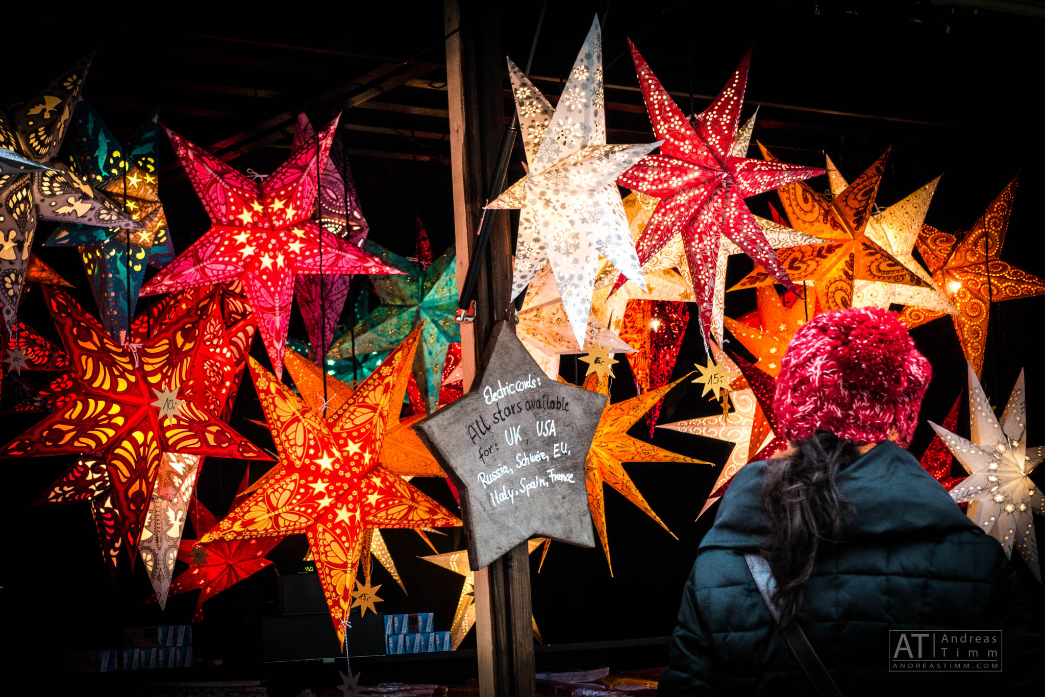 A person wearing a red knit hat admires vibrant, illuminated star decorations at a festive market stall.