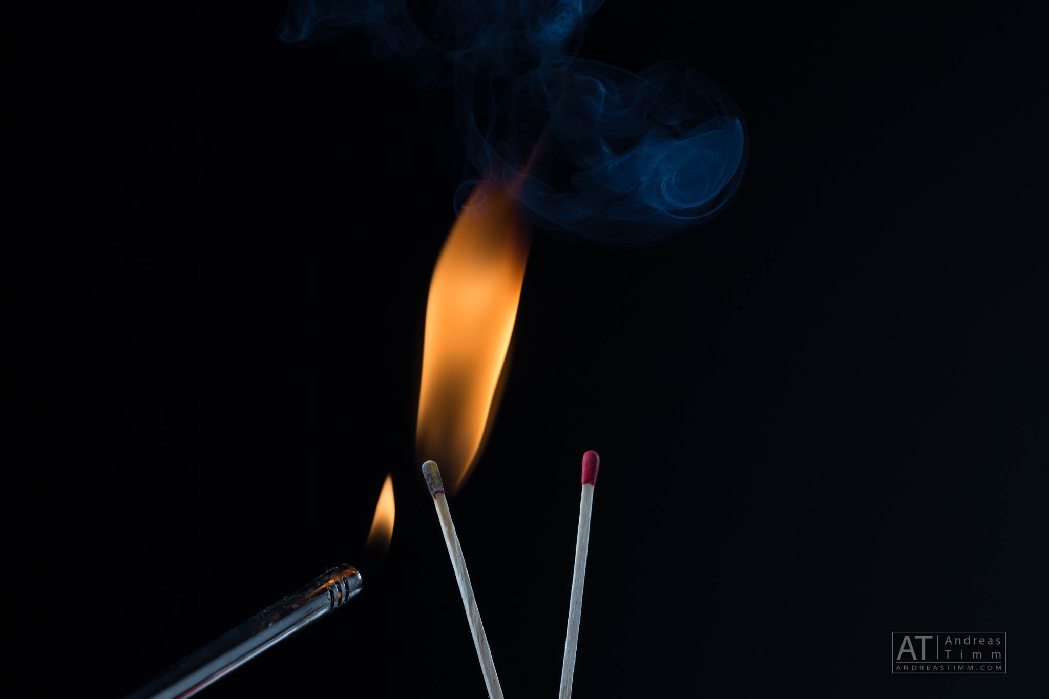 A lit match emits a bright flame and smoke against a dark background, with a lighter nearby.