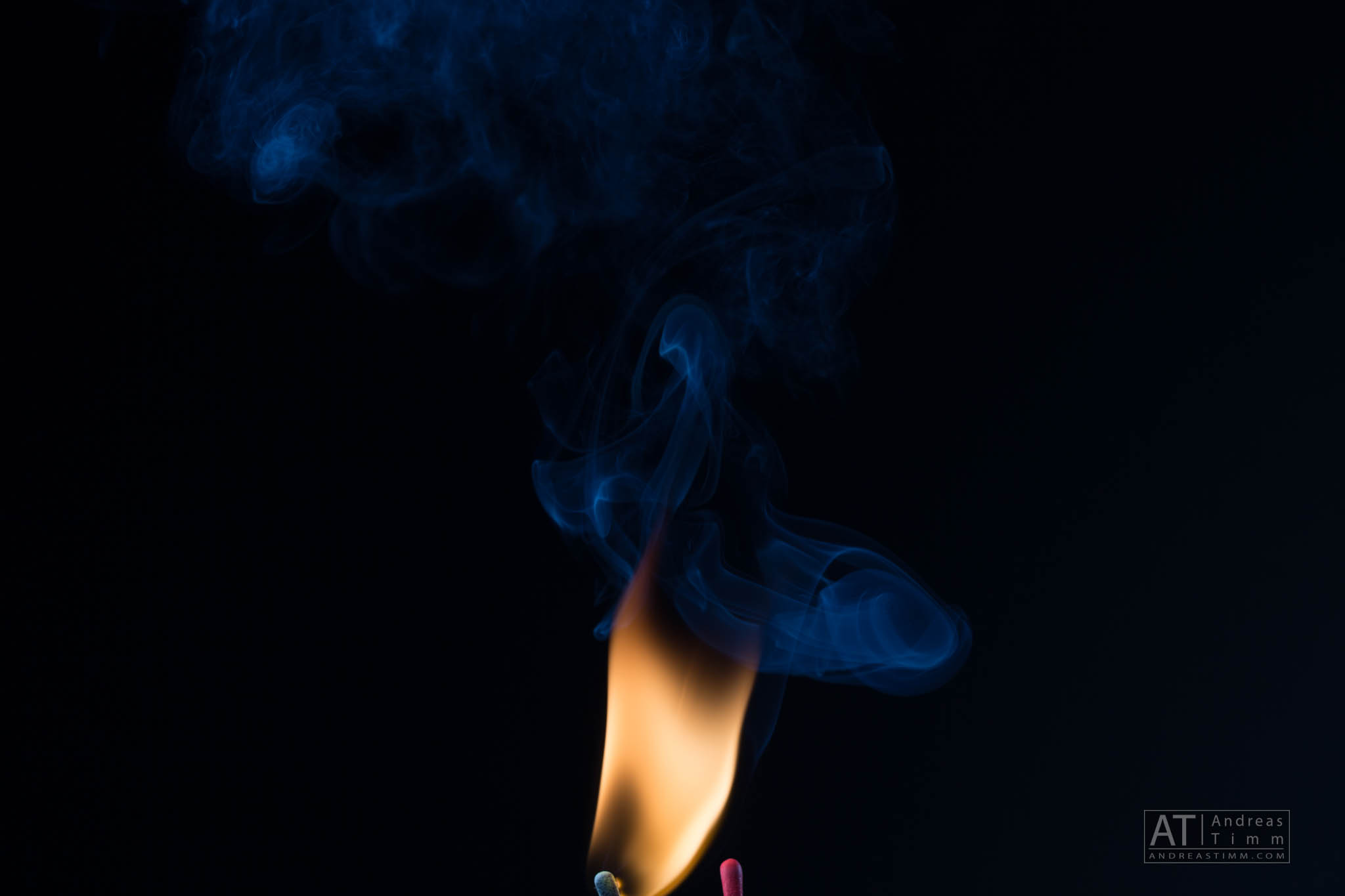 Blue smoke swirls around a lit match in the dark, creating a striking contrast against the black background.