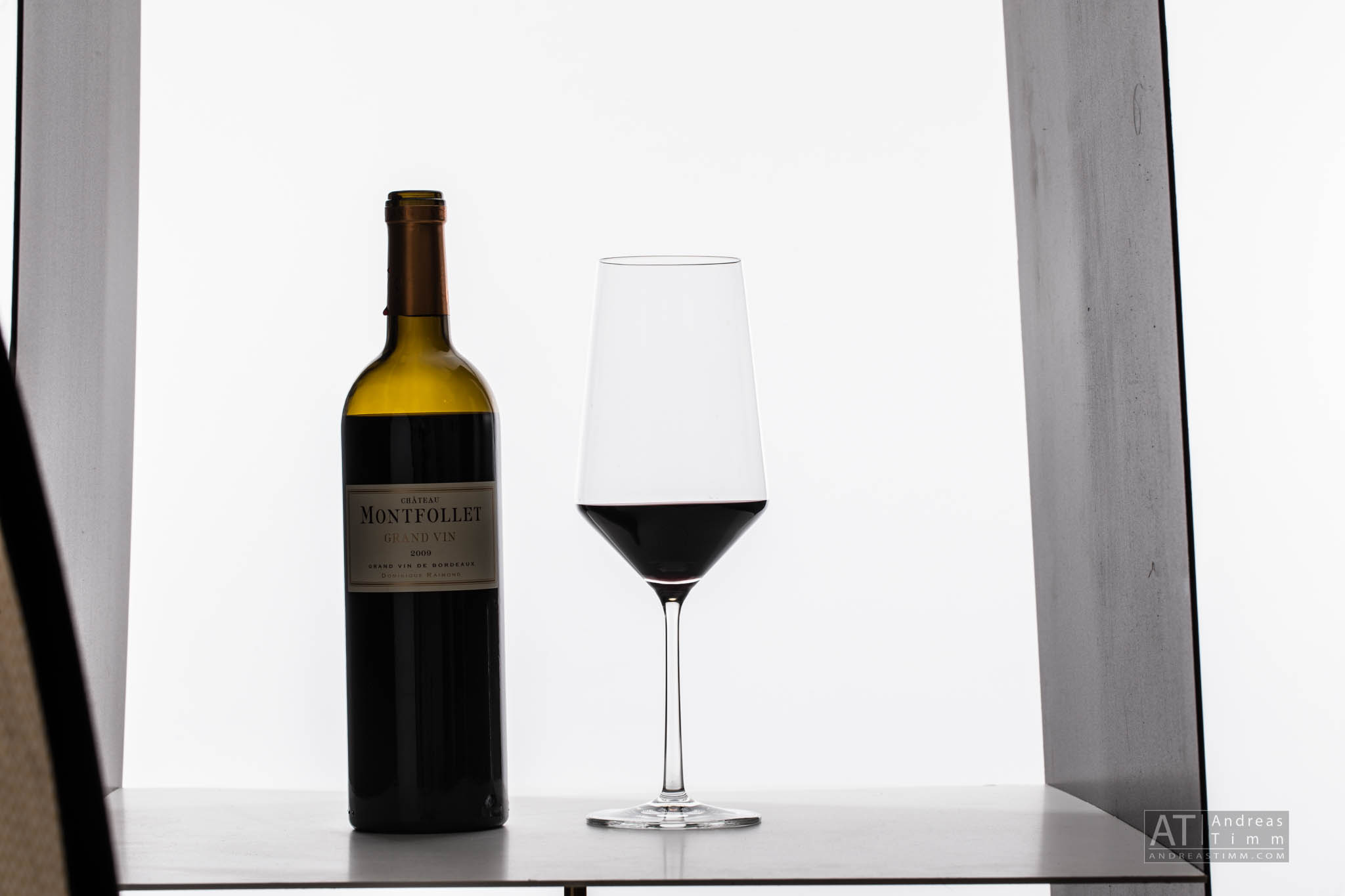 Bottle of Château Montfollet wine next to a filled wine glass on a light background.