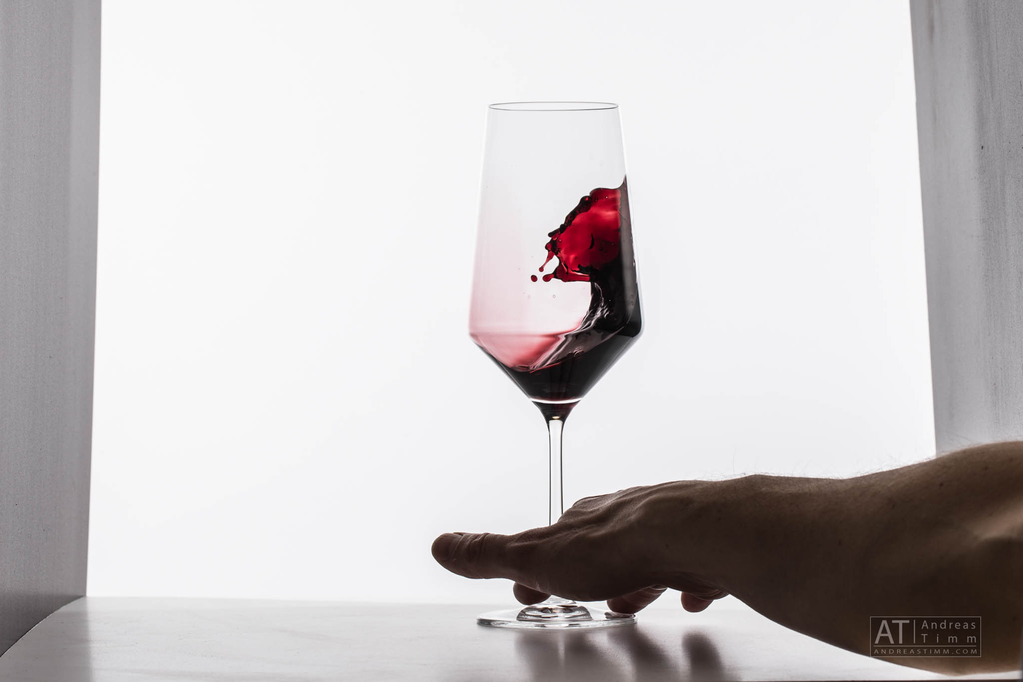 Hand tilting glass of red wine with a swirling splash on a white background.
