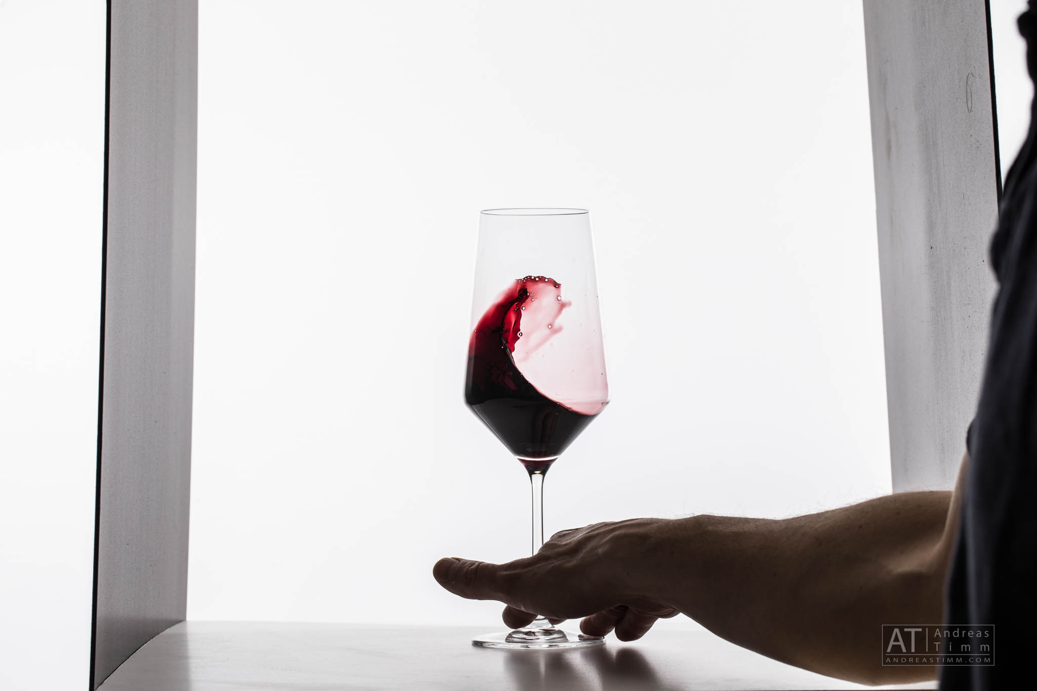 Hand holding stem of wine glass with swirling red wine against white background.
