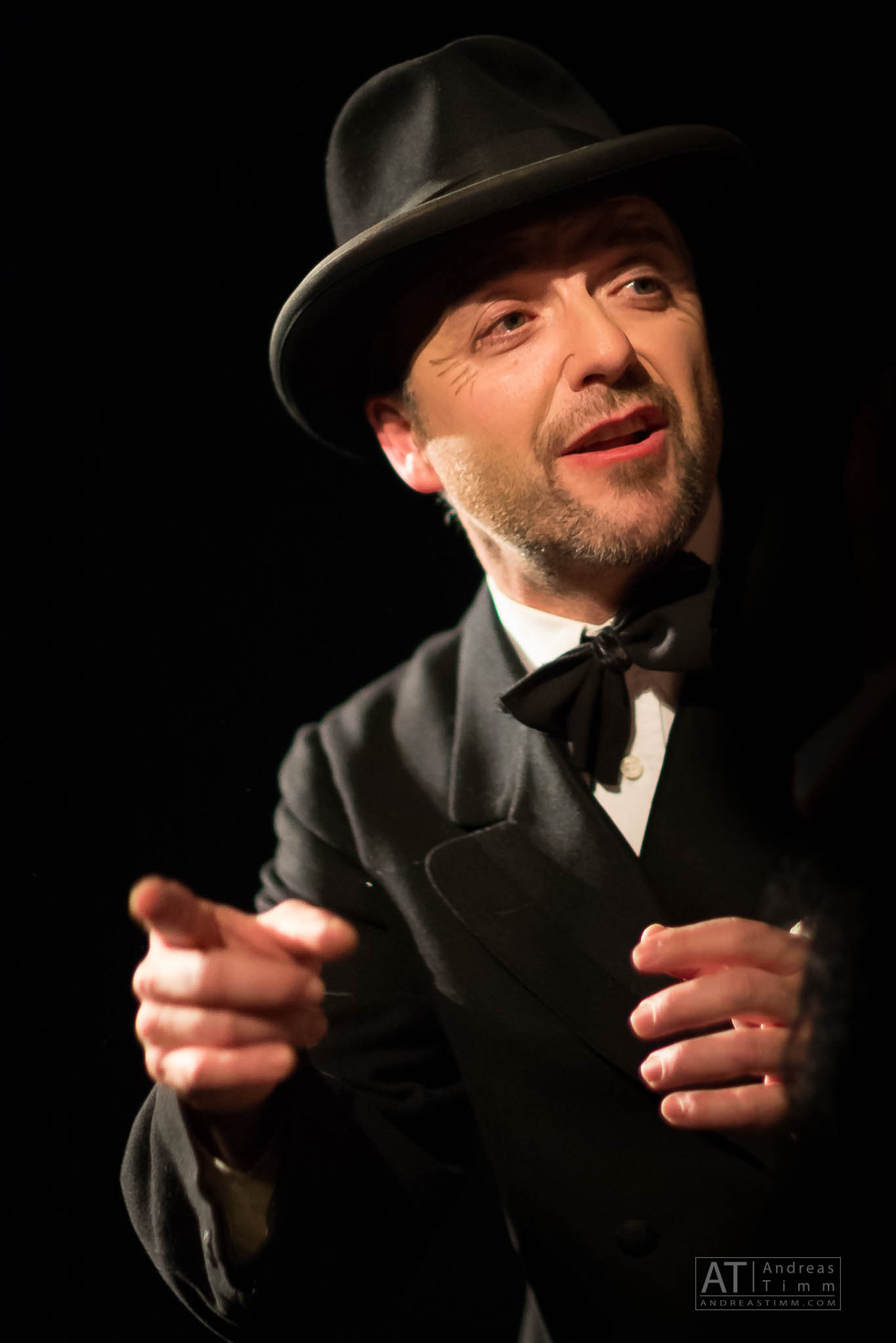 Man in vintage suit and bowler hat gesturing expressively on stage.