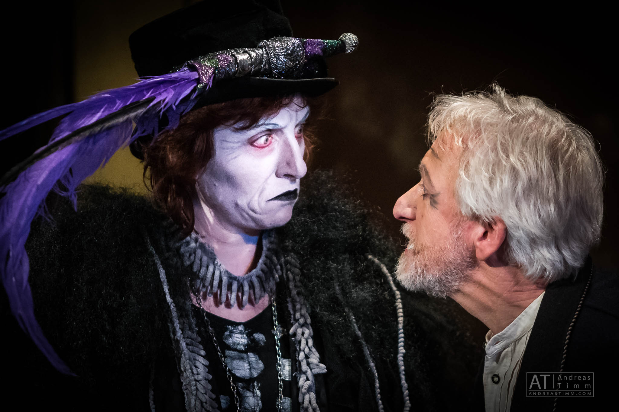 Two actors in dramatic costumes engage intensely, one with white face paint and purple feathers, the other with a white beard.