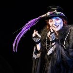 Performer in a black costume with a purple feathered hat, dramatic makeup, and striped gloves on a dark stage.