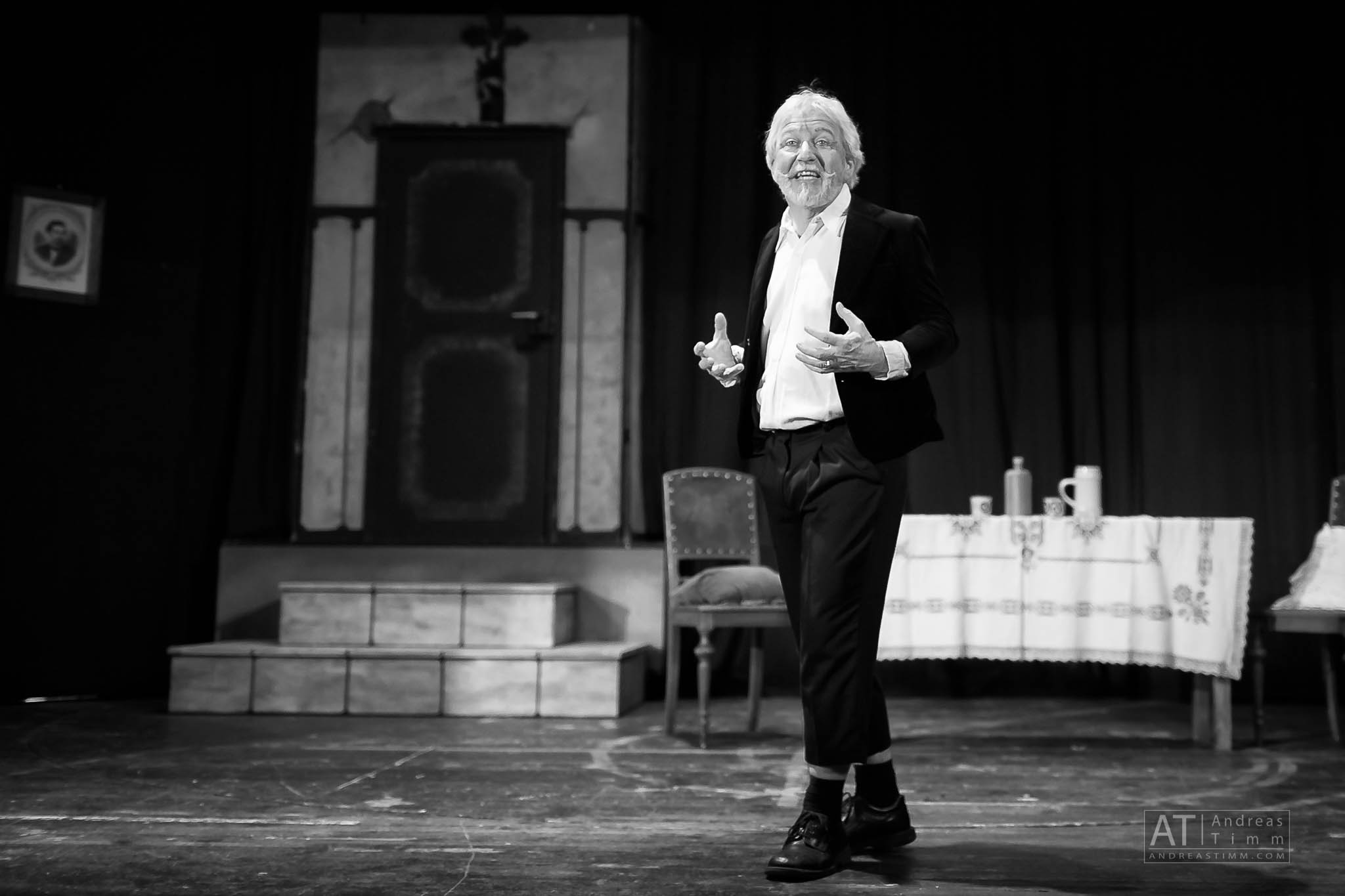 Actor performing energetically on a theater stage, monochrome setting with vintage decor and table.