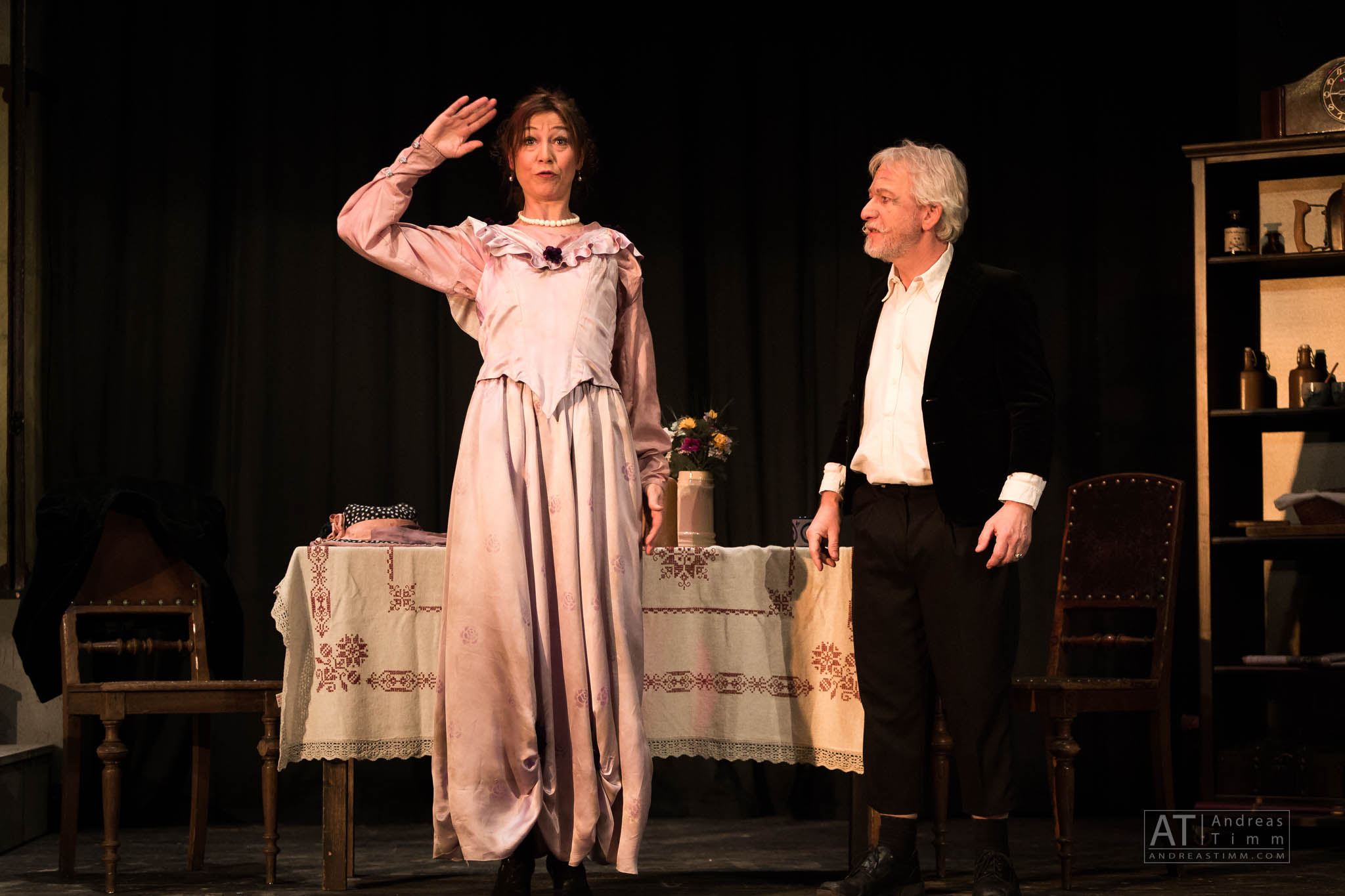 Two actors perform on stage, one in a pink vintage dress, the other in a black jacket, with a table and chairs behind them.