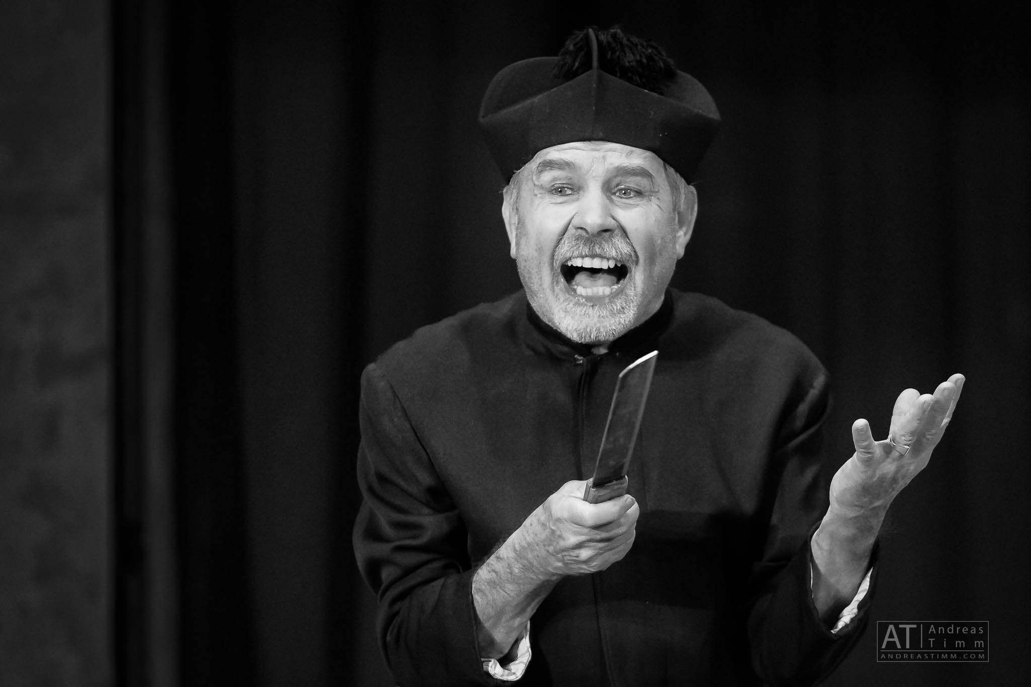 Black and white image of an enthusiastic man in a hat holding a knife, performing on stage.