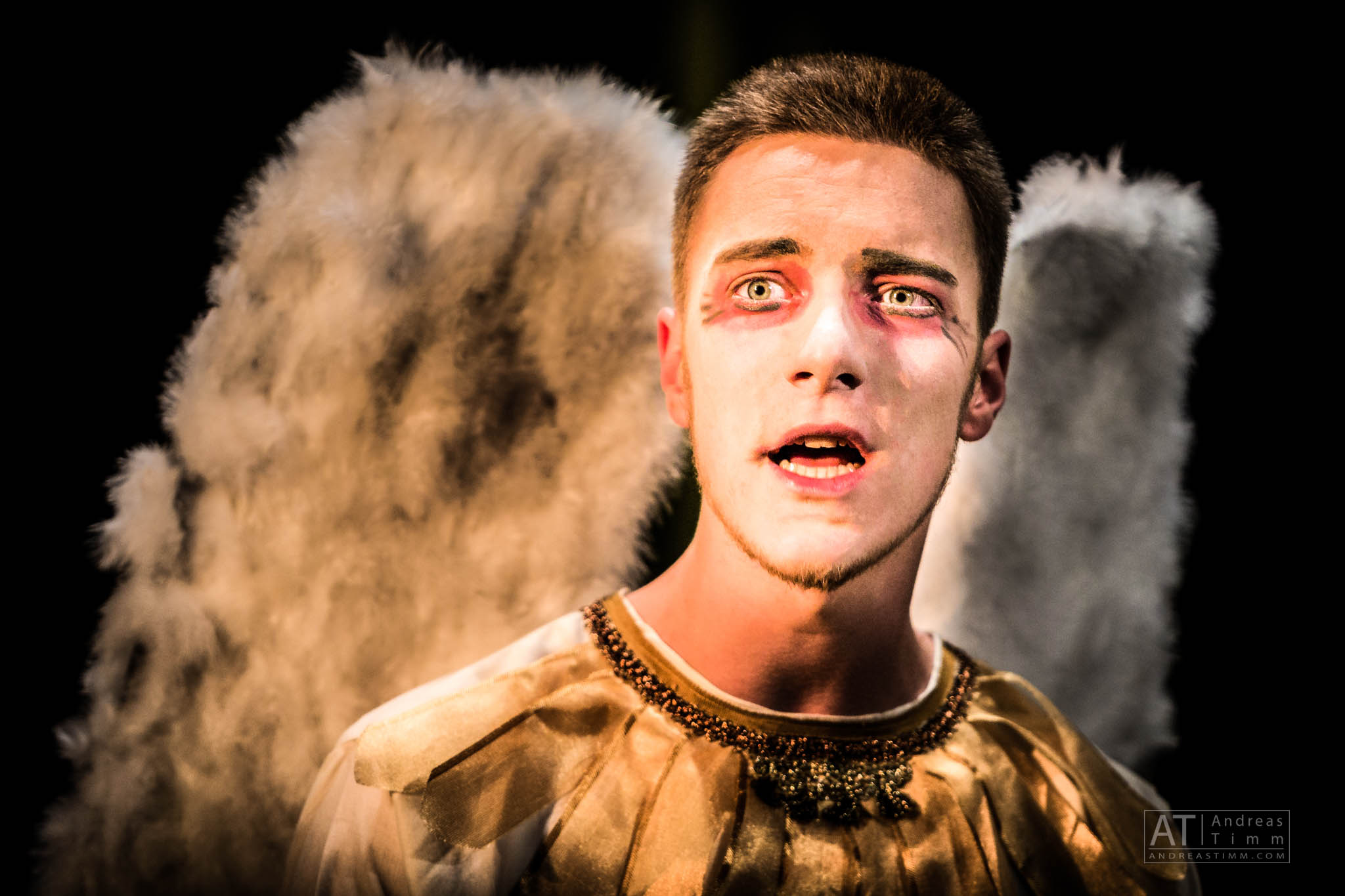 Actor in angel costume with dramatic makeup and white wings, mouth open as if speaking or singing, on stage.