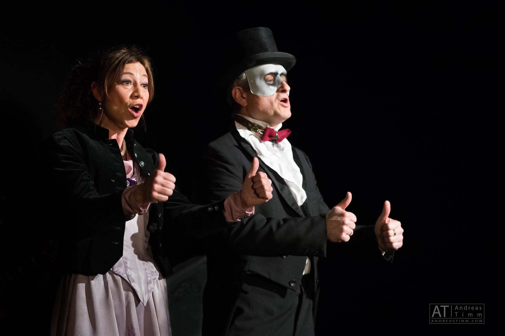 Two performers in theatrical costumes giving thumbs up on stage, man in top hat with face paint.