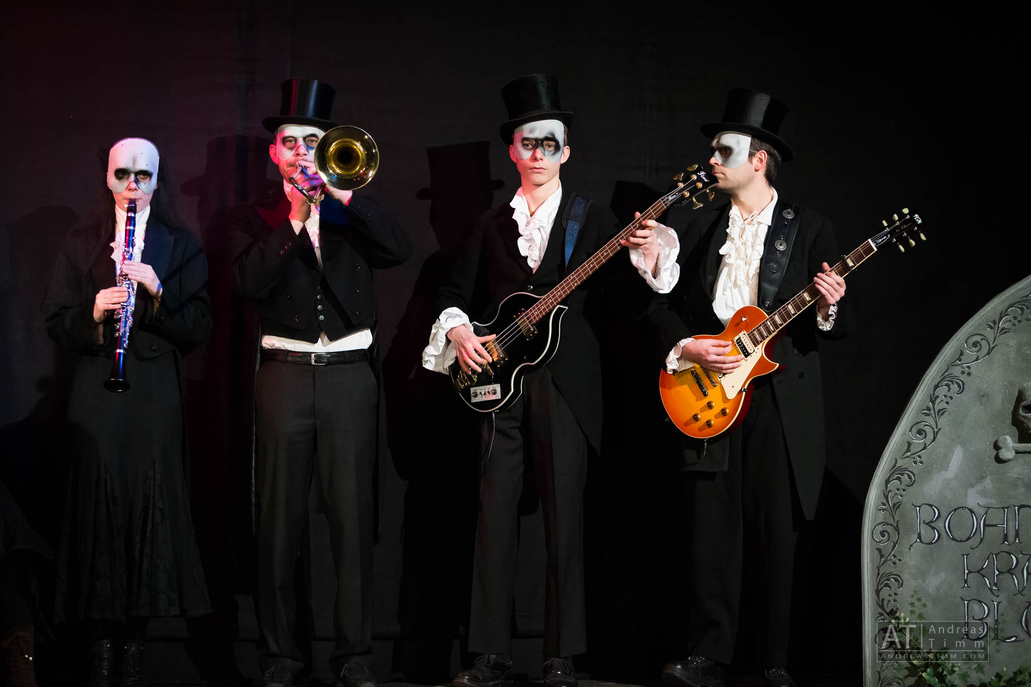 Band on stage in dark costumes and face paint, playing clarinet, trombone, bass, and guitar in a theatrical setting.