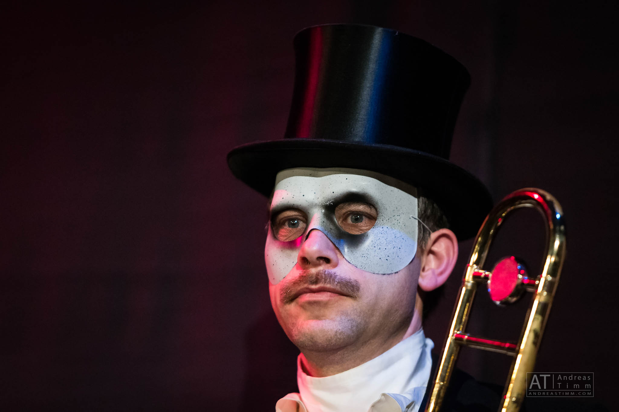 Man in top hat with white eye mask holding a trombone, dramatic costume on stage.