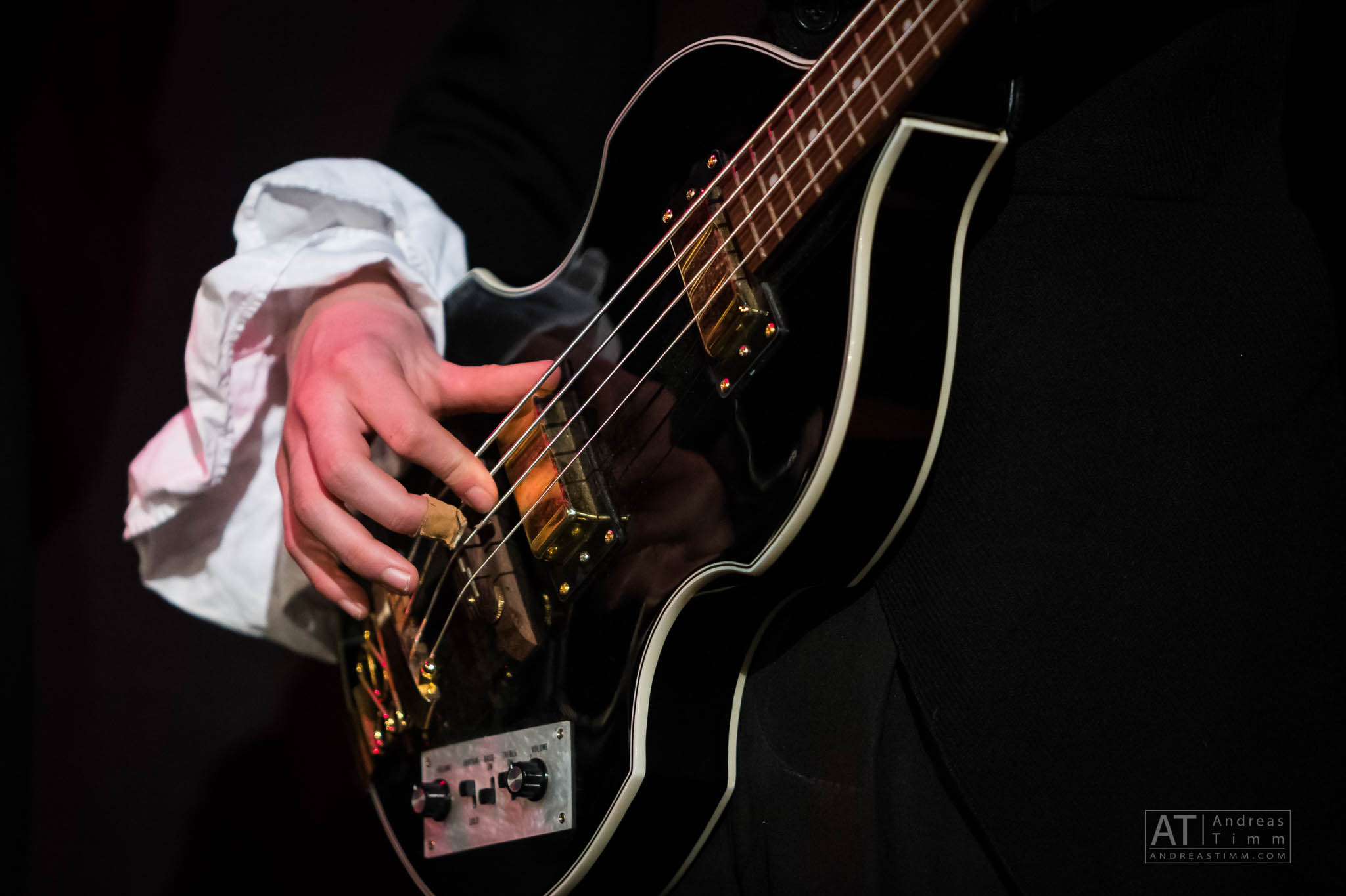 Close-up of a hand playing a black electric bass guitar with a gold bridge under stage lighting.