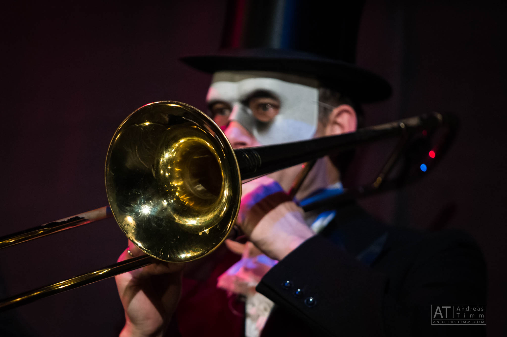 Musician playing trombone, wearing a top hat and half-mask in a dark, theatrical setting.