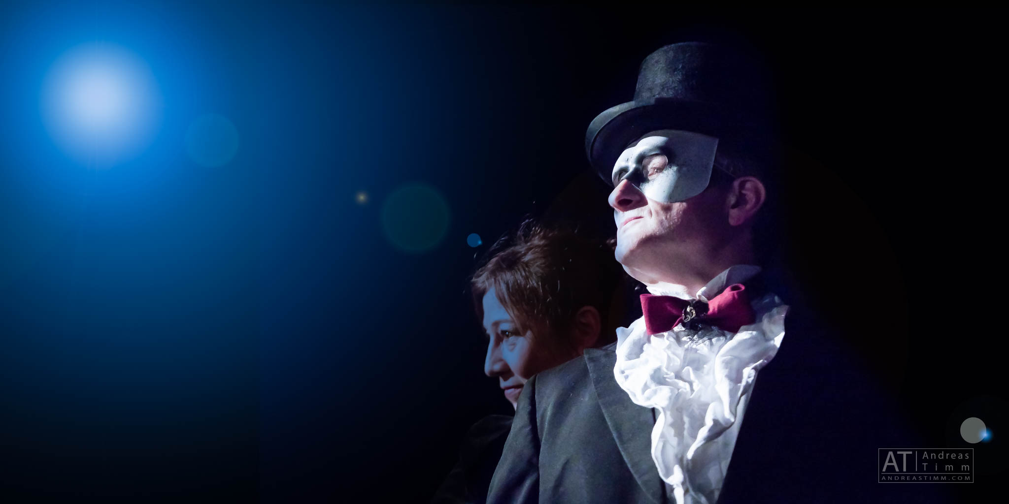 Man in a top hat and mask with a woman beside him under dramatic stage lighting.