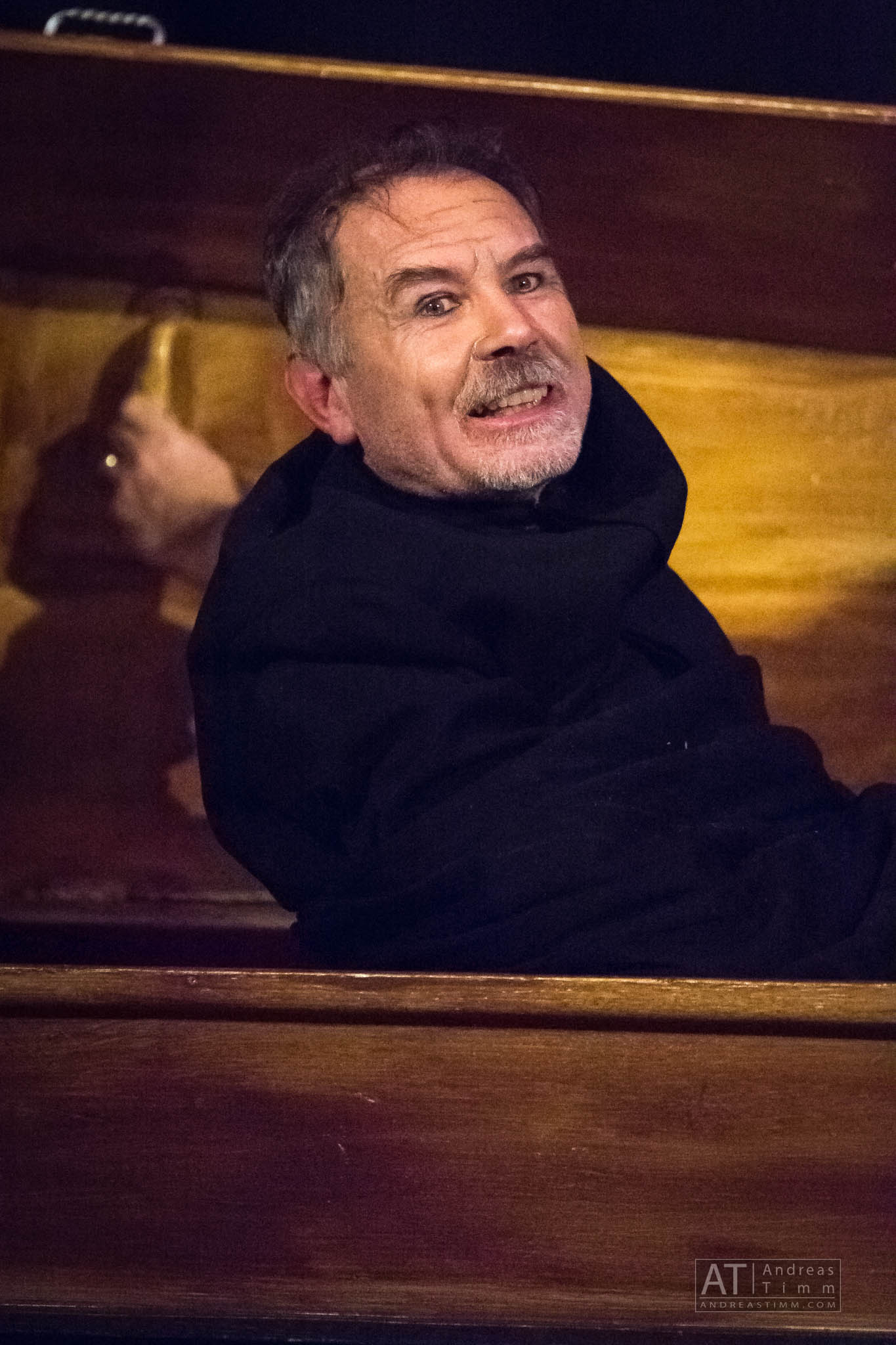 Man in dark robe grinning hard, seated in a wooden structure, with a blurred hand on the left.